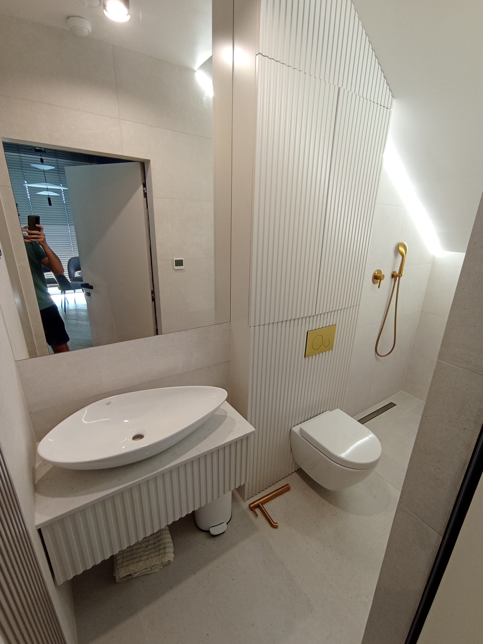 Nowoczesna łazienka w jasnych odcieniach z ryflowanymi szafkami, owalną umywalką nablatową, złotą armaturą i widocznym odbiciem osoby robiącej zdjęcie w lustrze.