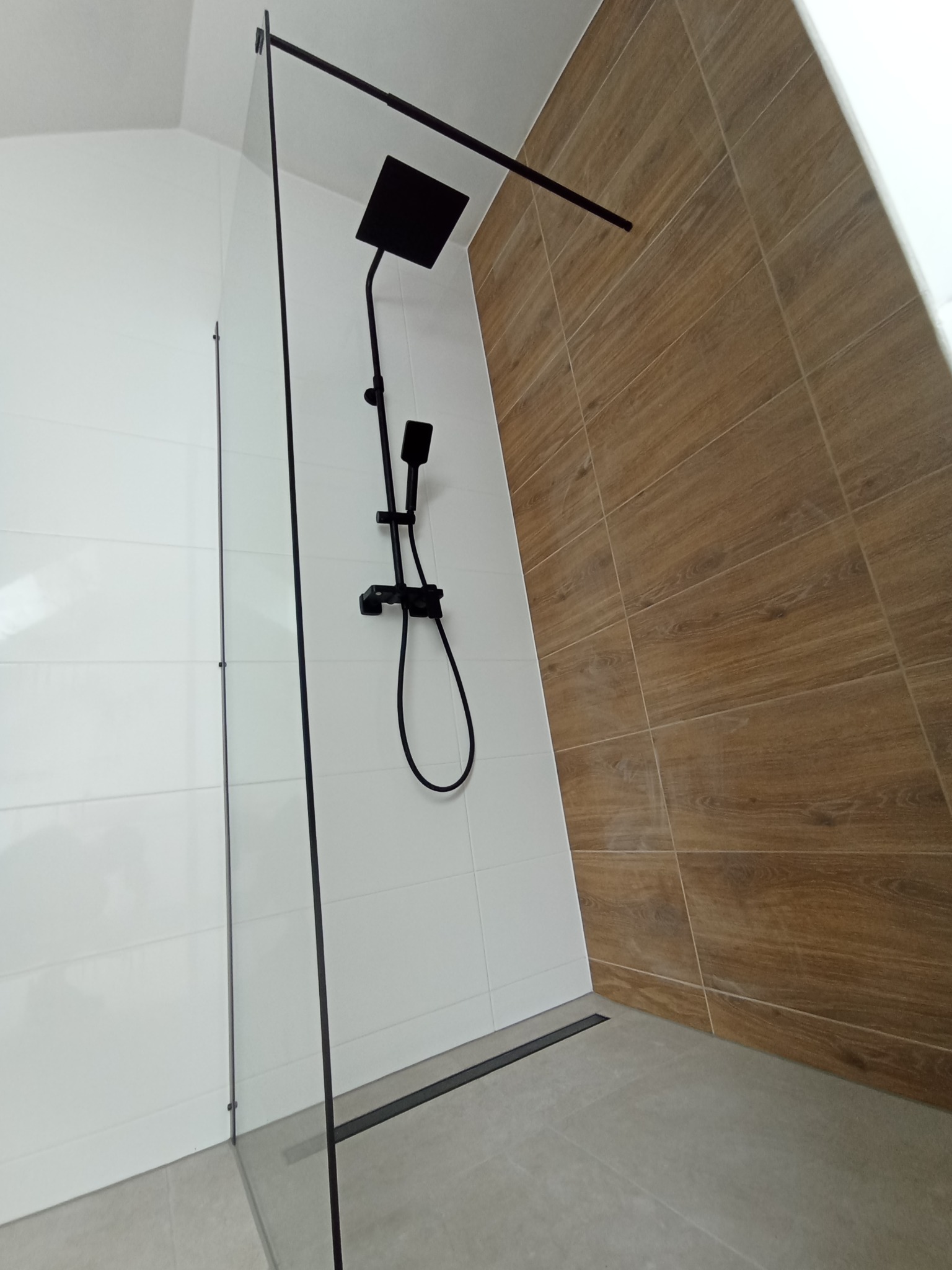 Nowoczesna kabina prysznicowa walk-in z czarną armaturą, odpływem liniowym i kombinacją białych i drewnopodobnych płytek na ścianach.