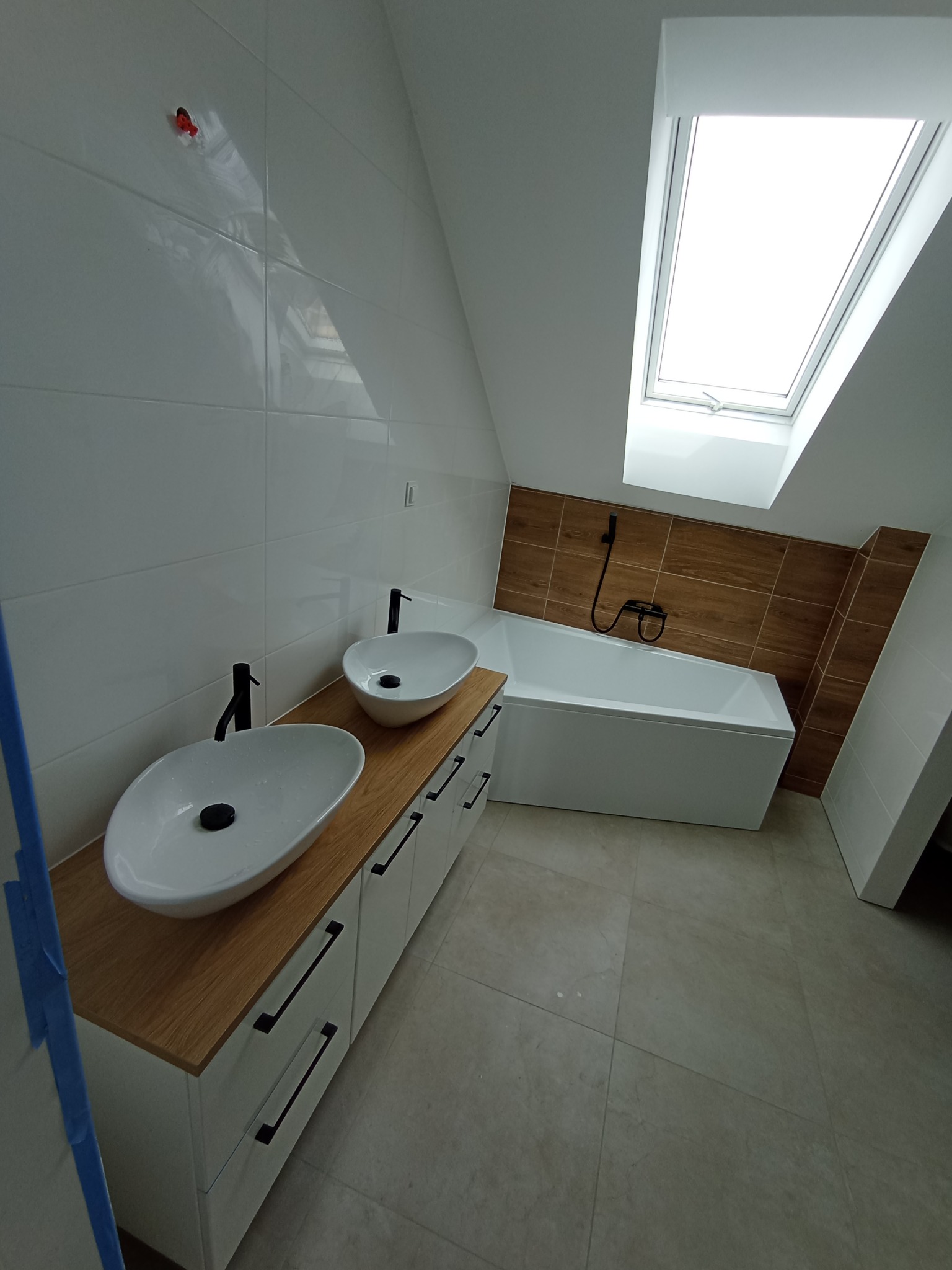 Nowoczesna łazienka na poddaszu z dwoma owalnymi umywalkami nablatowymi z czarną armaturą, białą zabudową szafkową z poziomymi uchwytami, wanną obudowaną płytkami imitującymi drewno i oknem dachowym.