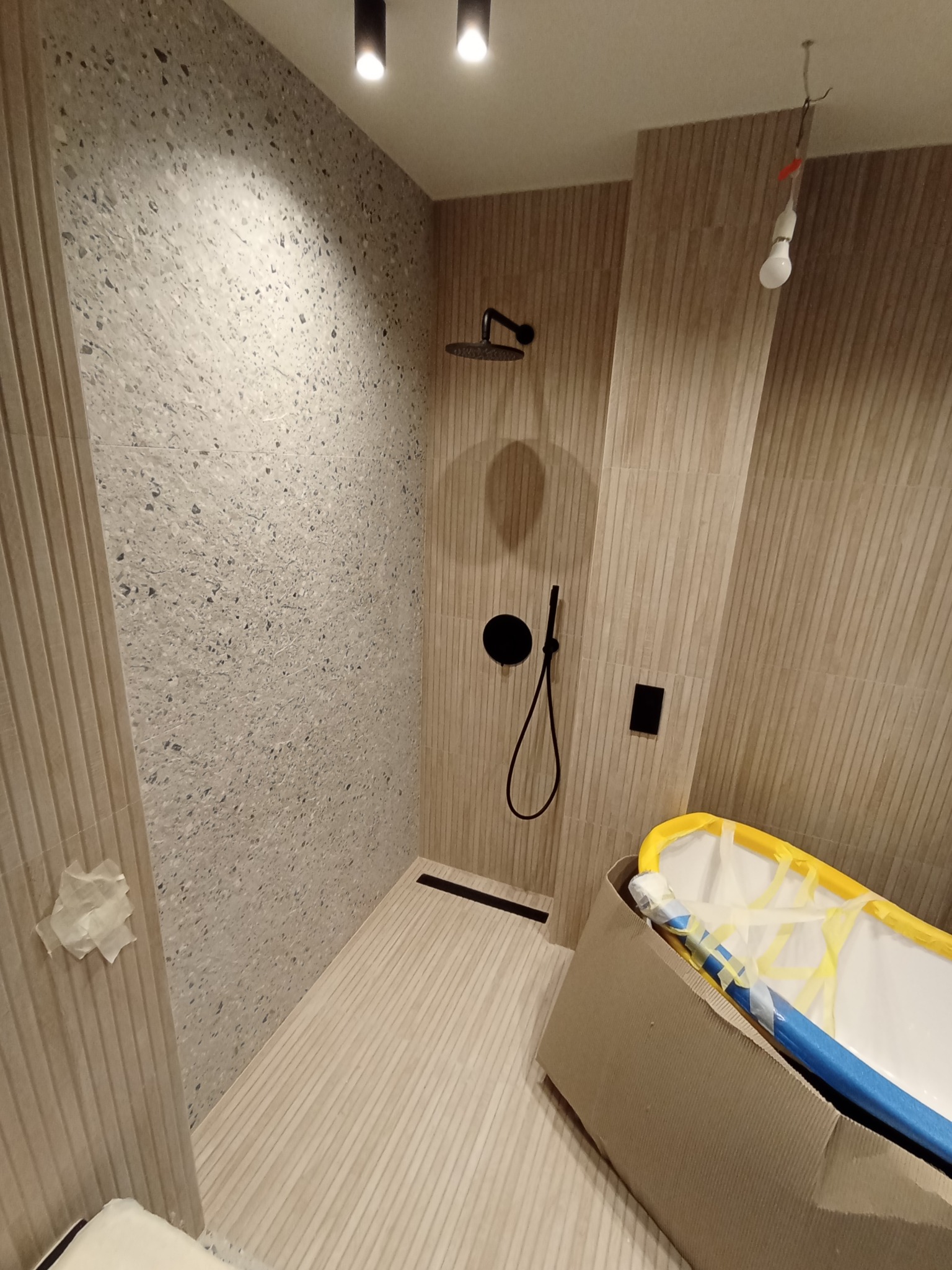 Nowoczesna kabina prysznicowa z deszczownicą, wykończona płytkami imitującymi drewno i kamień lastryko, z widoczną armaturą w kolorze czarnym i fragmentem wanny w kartonowym opakowaniu.