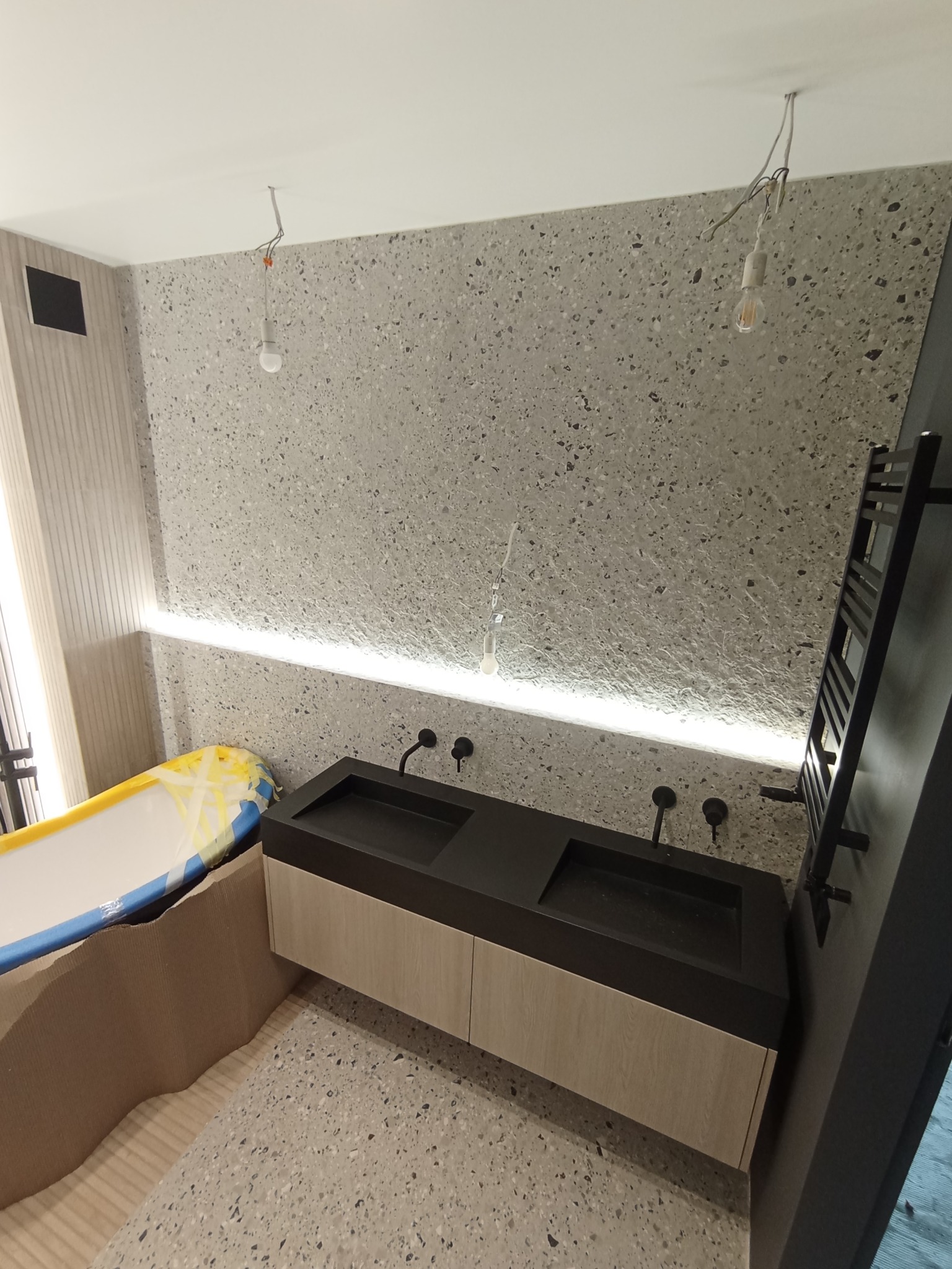 Podwójna umywalka z czarnym blatem i drewnopodobną szafką w łazience z terazzo, zabezpieczona wanna i niedokończone oświetlenie sufitowe.