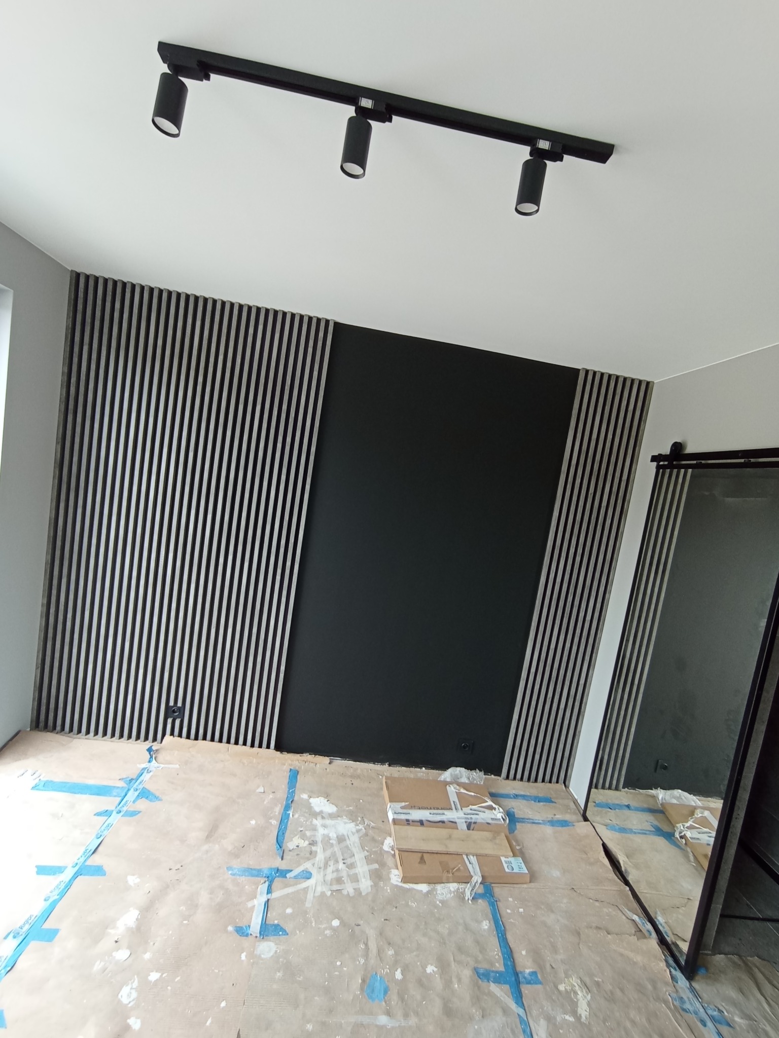 Montaż paneli lamelowych na ścianie w trakcie prac wykończeniowych, zabezpieczona podłoga, czarna ściana w tle, widoczne oświetlenie sufitowe.