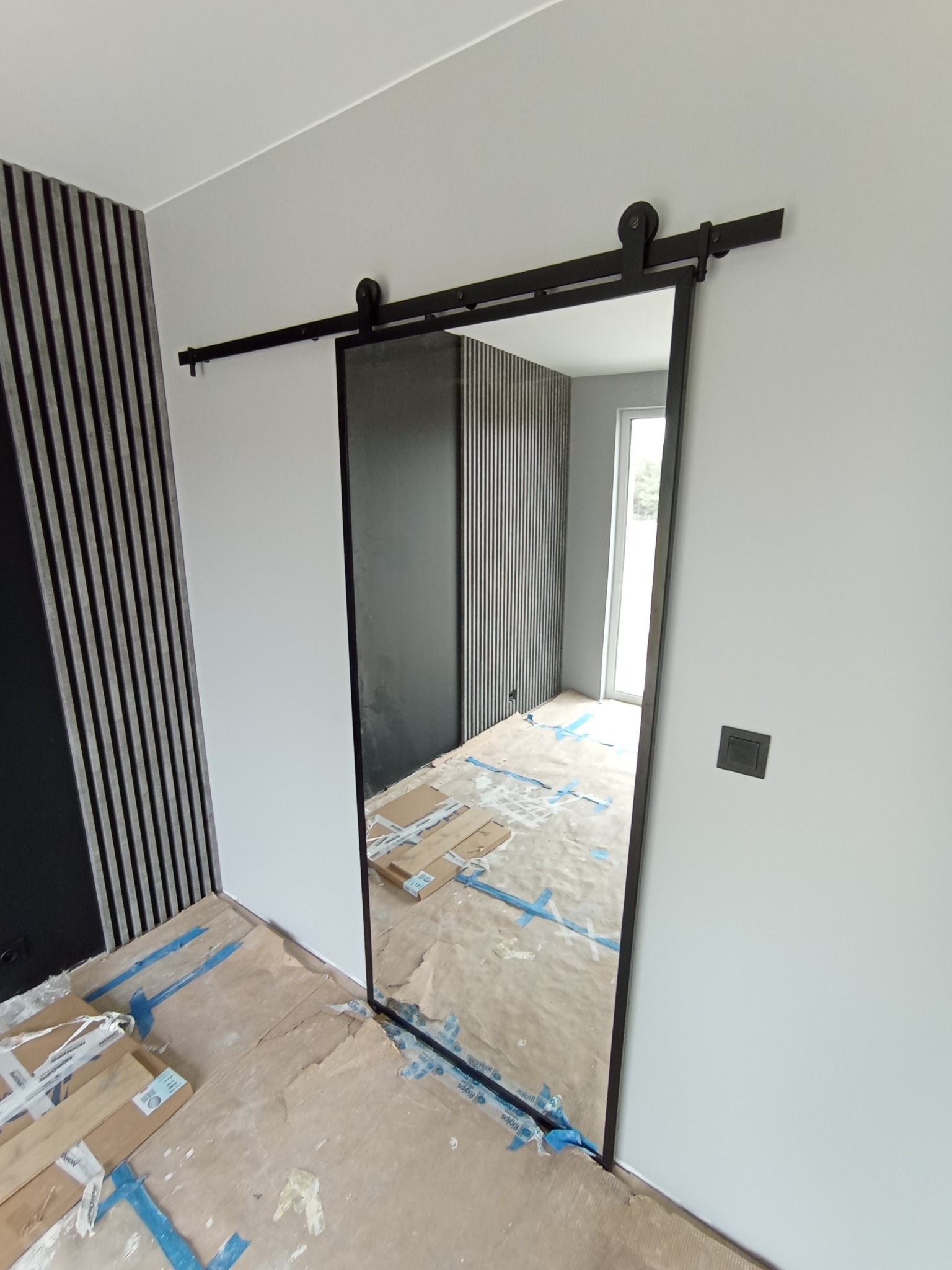 Montaż przesuwnych drzwi z czarną ramą i szyną w pomieszczeniu z panelami ściennymi, widoczne zabezpieczenia podłogi i elementy wykończeniowe.