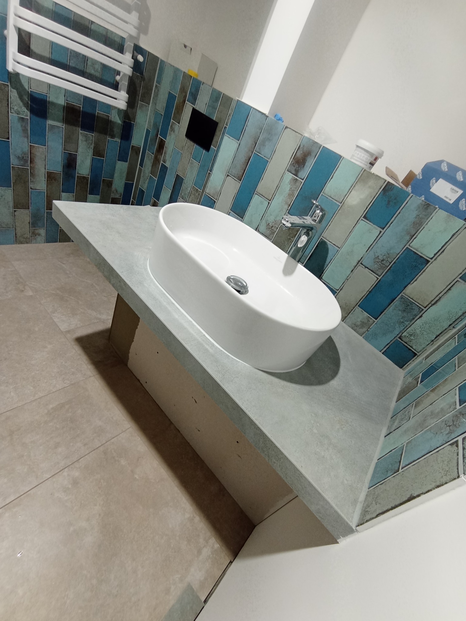 Łazienka w trakcie prac: owalna umywalka nablatowa na szarym blacie, niebiesko-szare płytki na ścianie ułożone w cegiełkę, widoczny grzejnik drabinkowy, pudełka i pojemniki w tle.