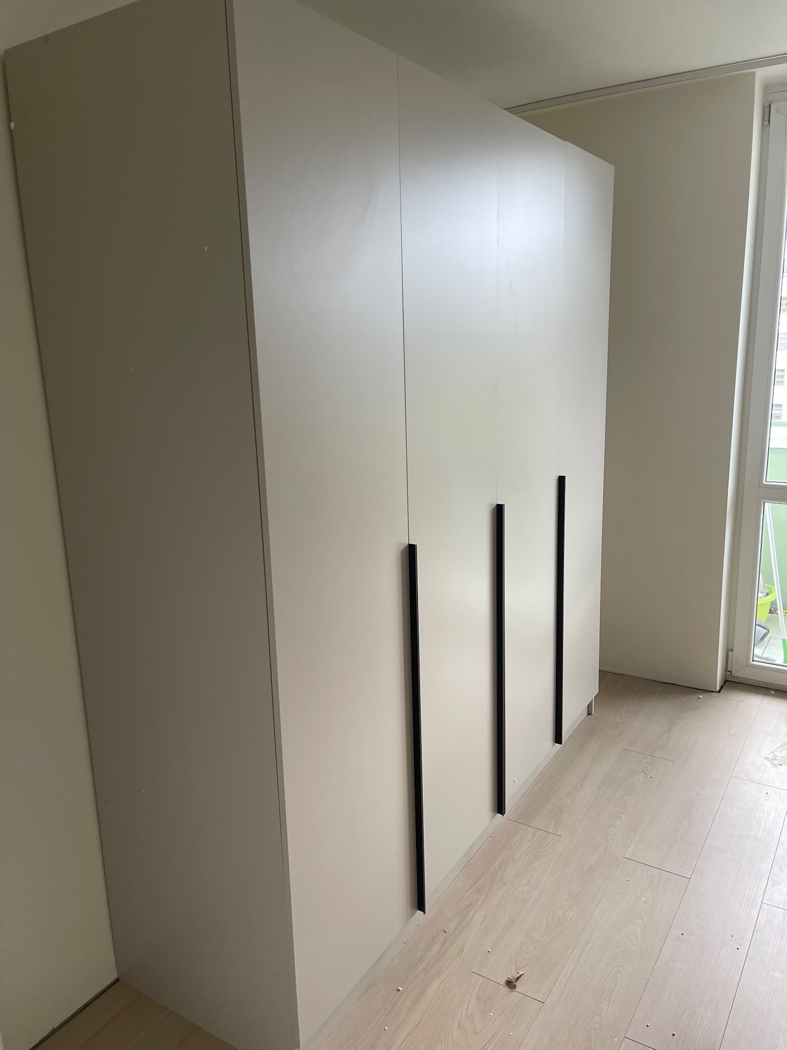 Jasny, trzydrzwiowy, minimalistyczny model szafy z podłużnymi, czarnymi uchwytami, ustawiony na panelach podłogowych w nowym wnętrzu.