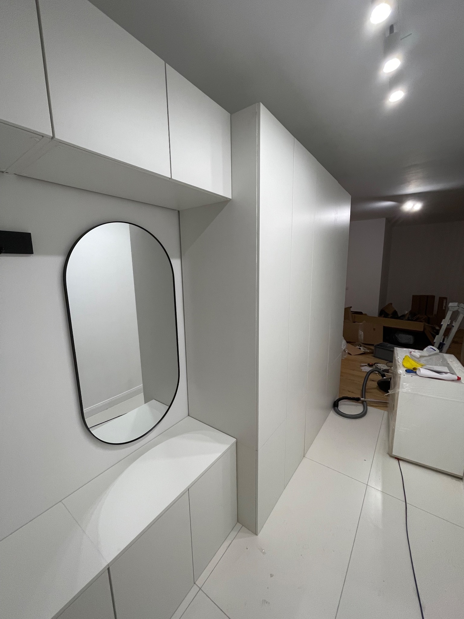 Biała zabudowa przedpokoju z owalnym lustrem i szafkami. Widoczne kartony i narzędzia sugerują trwający montaż. Minimalistyczny design, jasne oświetlenie.