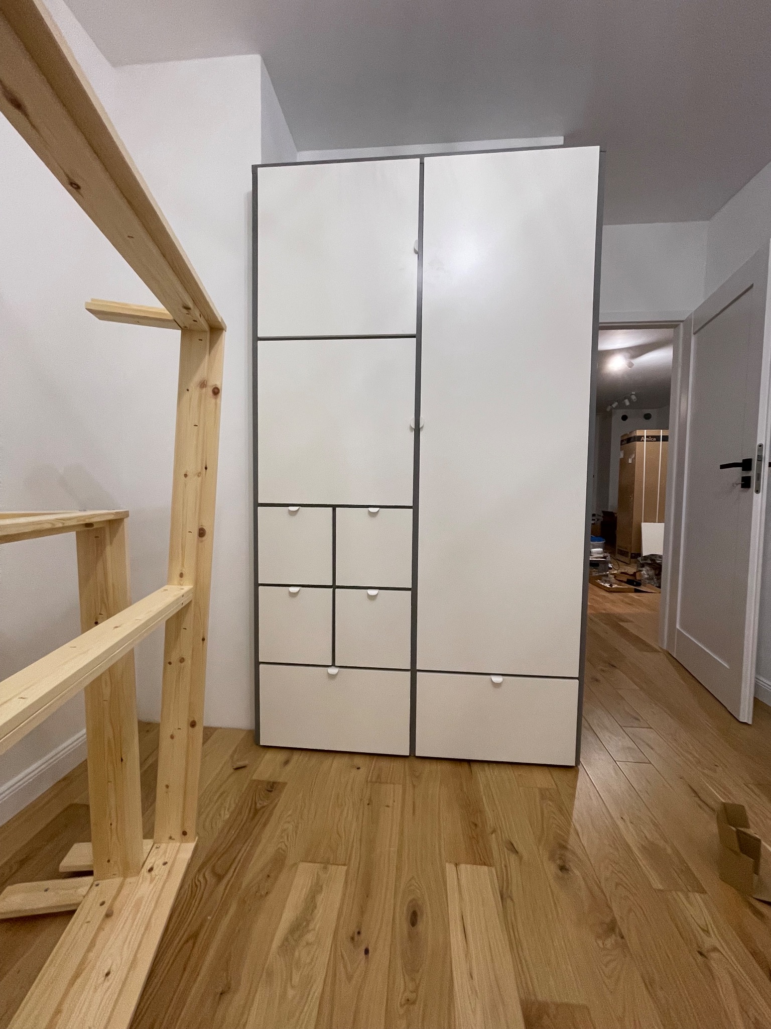 Biała szafa modułowa z szufladami i drewniana konstrukcja w jasnym wnętrzu. Parkiet na podłodze, otwarte drzwi w tle.