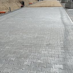 GRO-KOST - Ułożona szara kostka brukowa w jodełkę, z widocznym fragmentem przygotowanego podłoża piaskowego, rurami odprowadzającymi wodę i stosem kostki do układania w tle, w pobliżu budynku mieszkalnego...