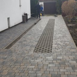 GRO-KOST - Ułożona szara kostka brukowa z centralnym pasem ażurowych elementów drenażowych, widoczny kanał odpływowy przy ścianie budynku i dwóch pracowników w tle.