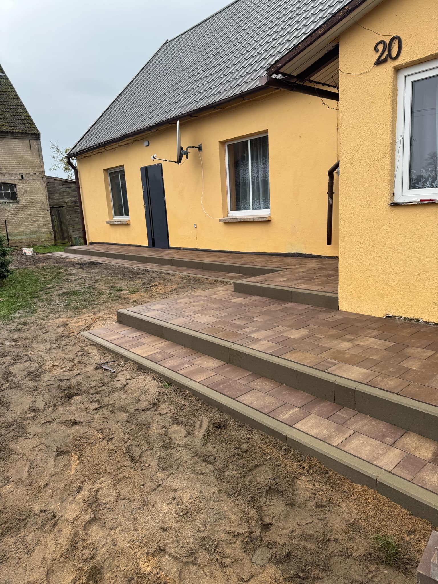Nowo ułożona, brązowa kostka brukowa tworząca schody i chodnik przed żółtym domem z numerem 20, widoczne ślady stóp w piasku.
