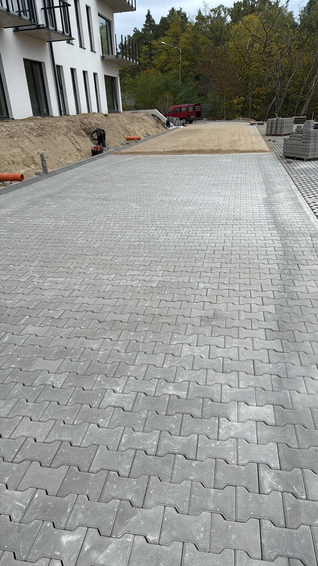 Ułożona szara kostka brukowa w jodełkę, z widocznym fragmentem przygotowanego podłoża piaskowego, rurami odprowadzającymi wodę i stosem kostki do układania w tle, w pobliżu budynku mieszkalnego...