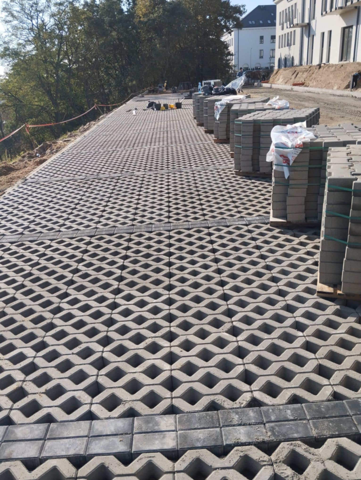 Szara, ażurowa nawierzchnia z paletami kostki po prawej stronie, w tle budynek i drzewa, widoczne ślady piasku między elementami.