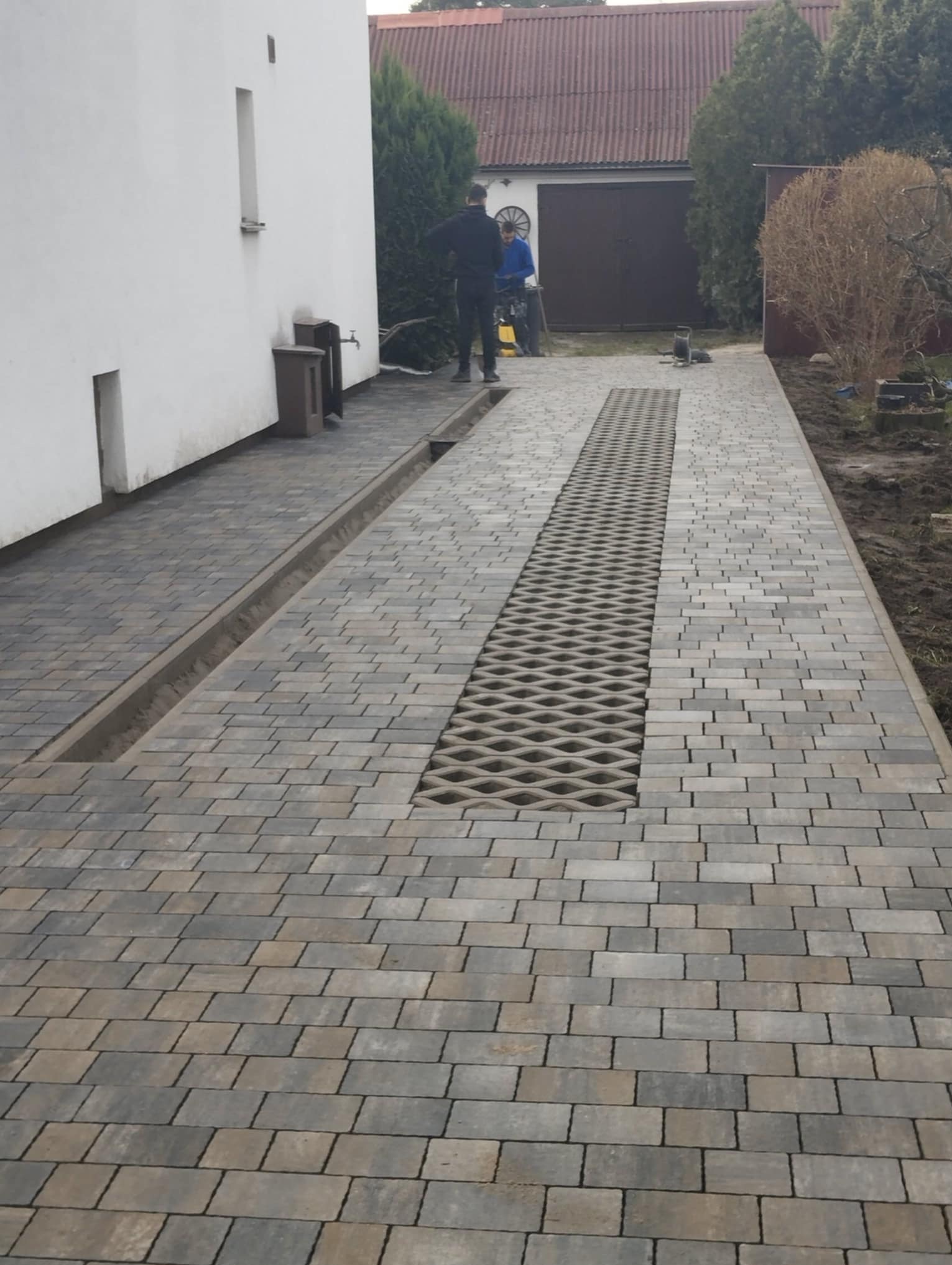 Ułożona szara kostka brukowa z centralnym pasem ażurowych elementów drenażowych, widoczny kanał odpływowy przy ścianie budynku i dwóch pracowników w tle.