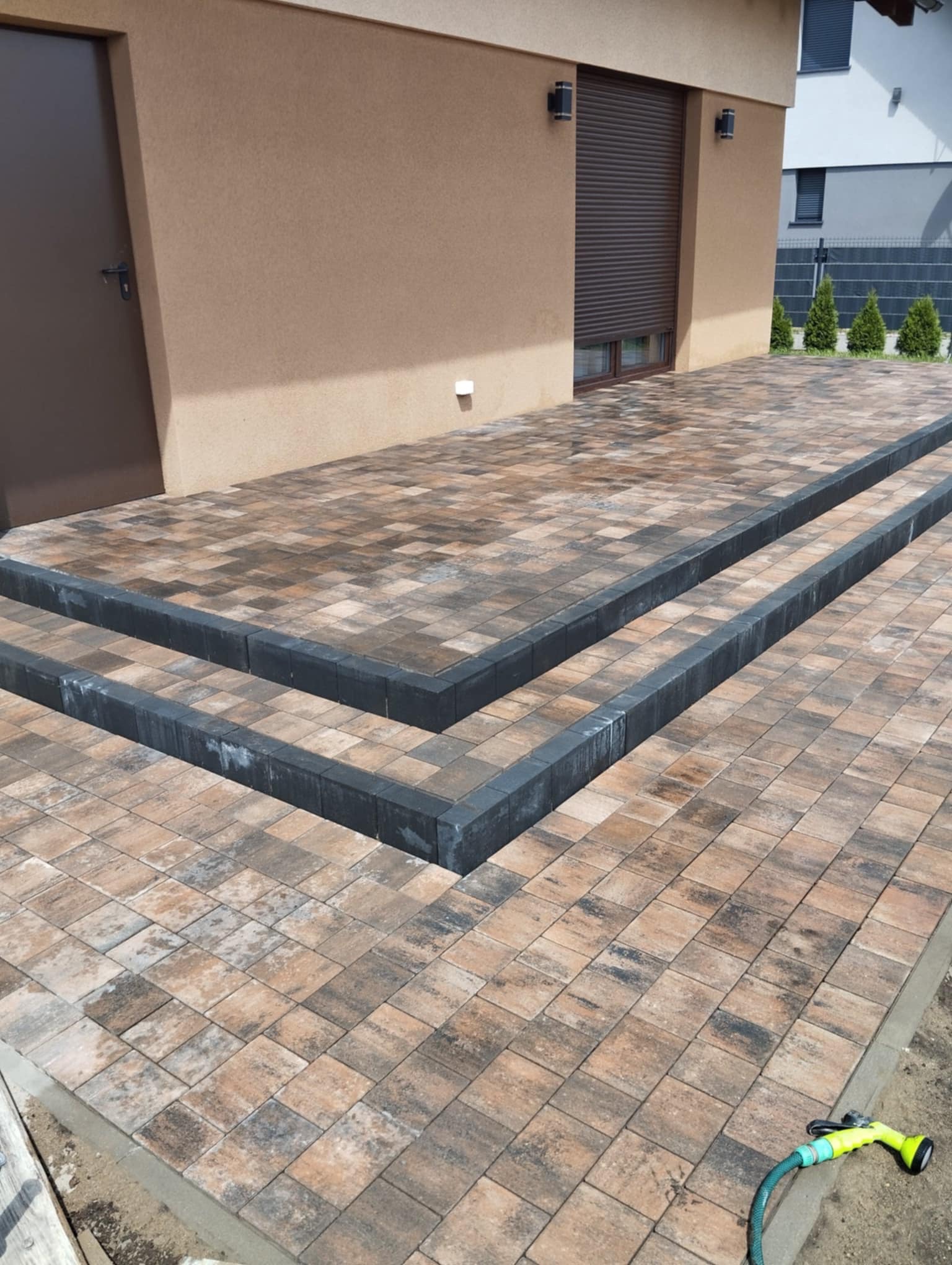 Ułożona kostka brukowa w odcieniach brązu i szarości tworząca schody przed budynkiem z brązowymi roletami i drzwiami. Widoczny wąż ogrodowy.