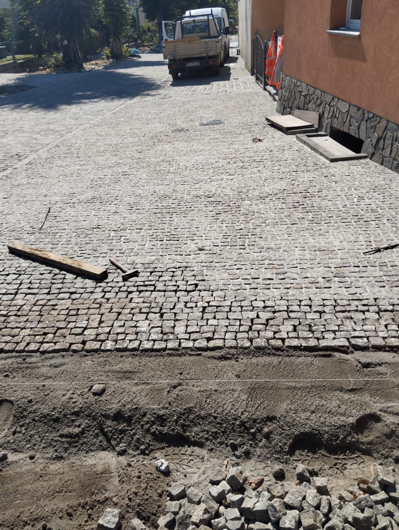 Układanie kostki granitowej: Widok na fragment placu z ułożoną kostką granitową, z widocznymi narzędziami (drewniana deska, młotek) i przygotowanym podłożem z piasku. W tle budynek i samochód...