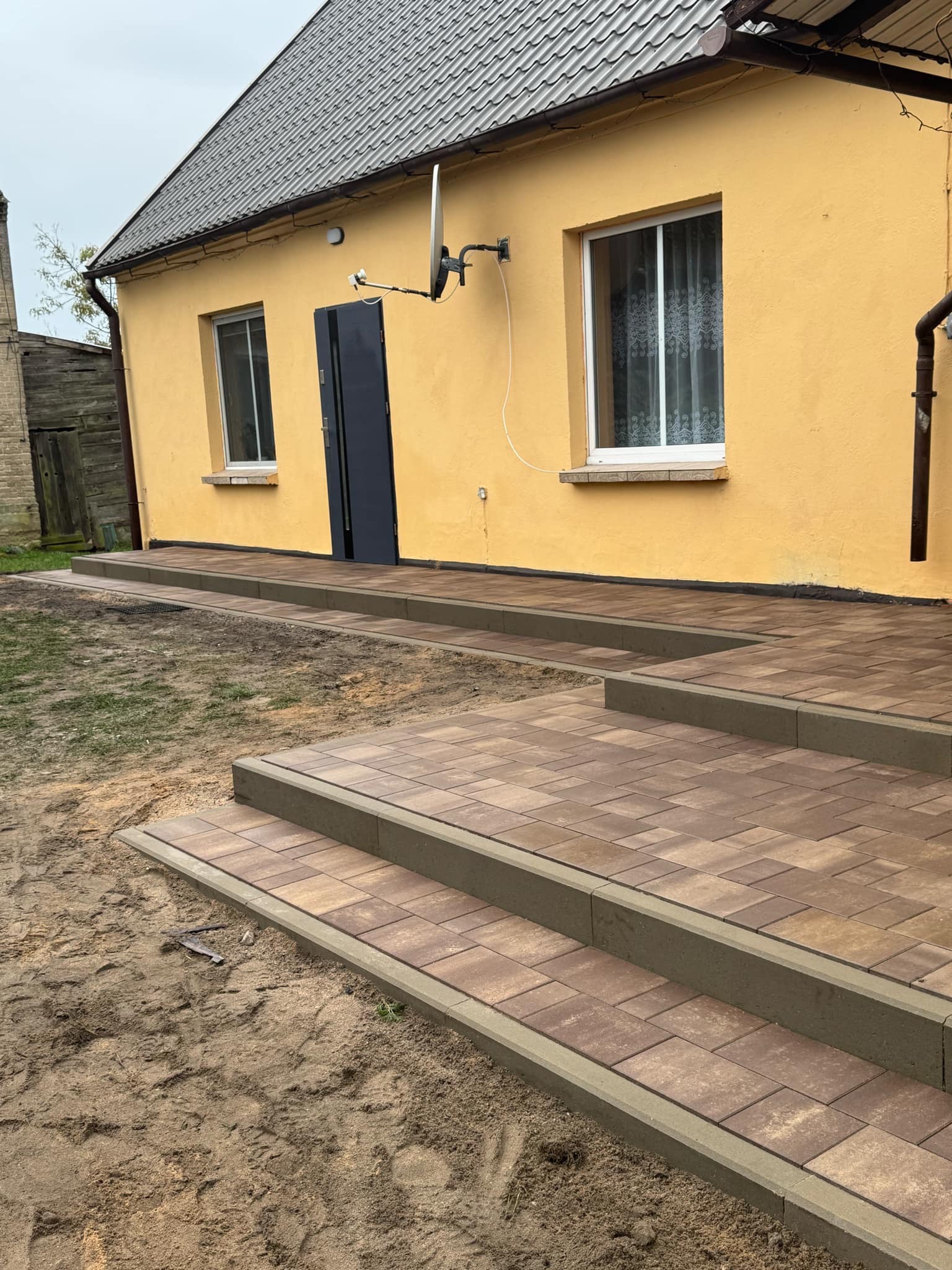 Ułożona kostka brukowa tworząca schody i chodnik przed żółtym domem z szarą dachówką, widoczne niedociągnięcia w postaci piasku na kostce.