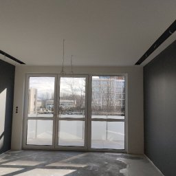 Dmytro Dutchak - Wnętrze w trakcie wykończenia: szare ściany, biały sufit z czarnymi listwami, duże okno balkonowe z widokiem na budynki, surowa podłoga przed położeniem paneli, wystające kable oświetleniowe.