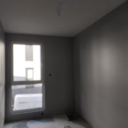 Dmytro Dutchak - Pokój po malowaniu na szaro, widoczne świeżo pomalowane ściany, okno z widokiem na budynek w tle, surowy stan podłogi.