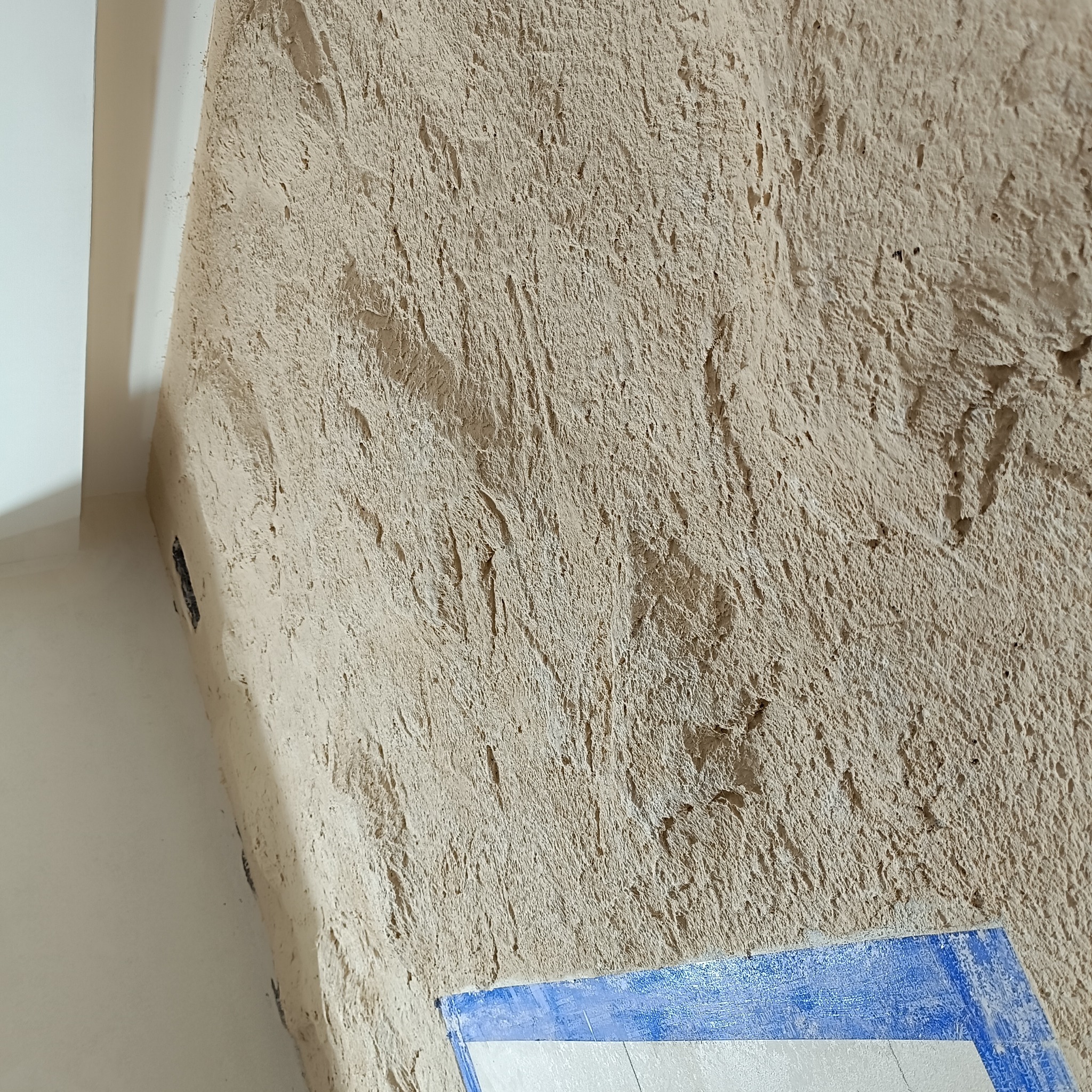 Szczegółowe ujęcie ściany z grubą fakturą nałożonej warstwy w Bielsko-Białej, widoczne nierówności i detale struktury. W rogu fragment okleiny w kolorze niebieskim.