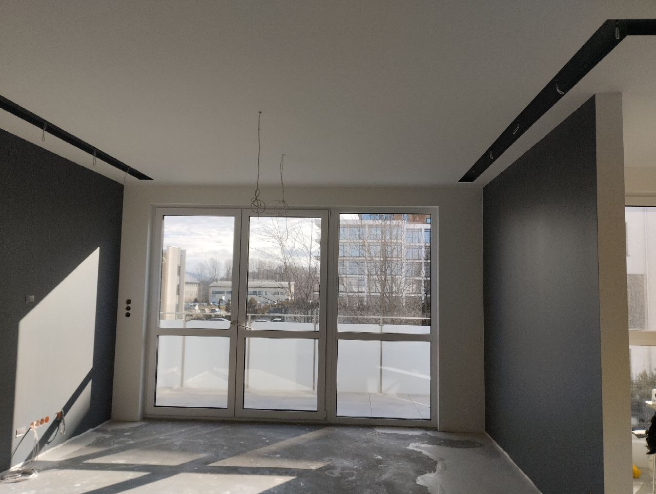 Wnętrze w trakcie wykończenia: szare ściany, biały sufit z czarnymi listwami, duże okno balkonowe z widokiem na budynki, surowa podłoga przed położeniem paneli, wystające kable oświetleniowe.