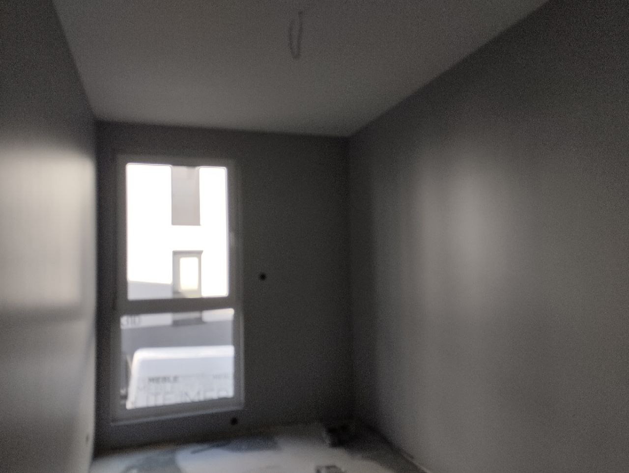 Pokój po malowaniu na szaro, widoczne świeżo pomalowane ściany, okno z widokiem na budynek w tle, surowy stan podłogi.