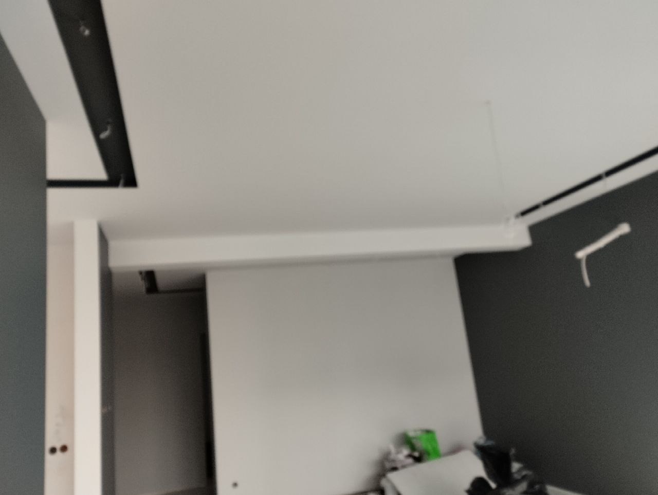 Wnętrze w trakcie wykańczania: pomalowane ściany w odcieniach szarości i bieli, widoczne prace przy suficie z elementami oświetlenia, na podłodze pozostałości po remoncie.