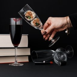 Piotr Kuchniak - Dwa kawałki sushi w kieliszku do szampana trzymanym przez kobiecą dłoń z brązowymi paznokciami i złotą bransoletką, obok kieliszka z czerwonym winem i odciskiem szminki, leżącego obok stosu książek...