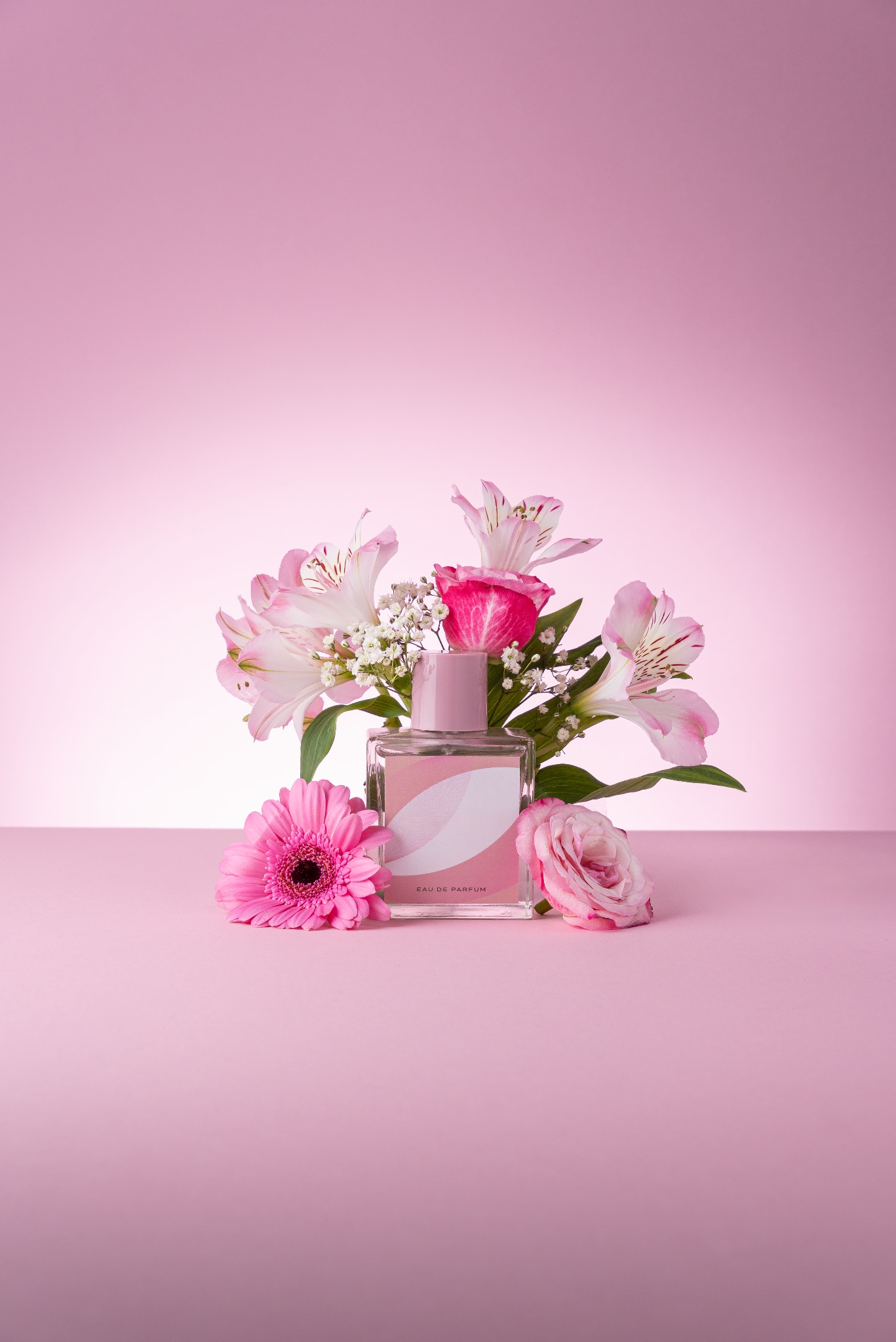 Szklany flakon perfum z różową etykietą, otoczony kompozycją z różowych kwiatów: gerbery, róży i alstremerii na tle jednolitego, pudrowego różu.