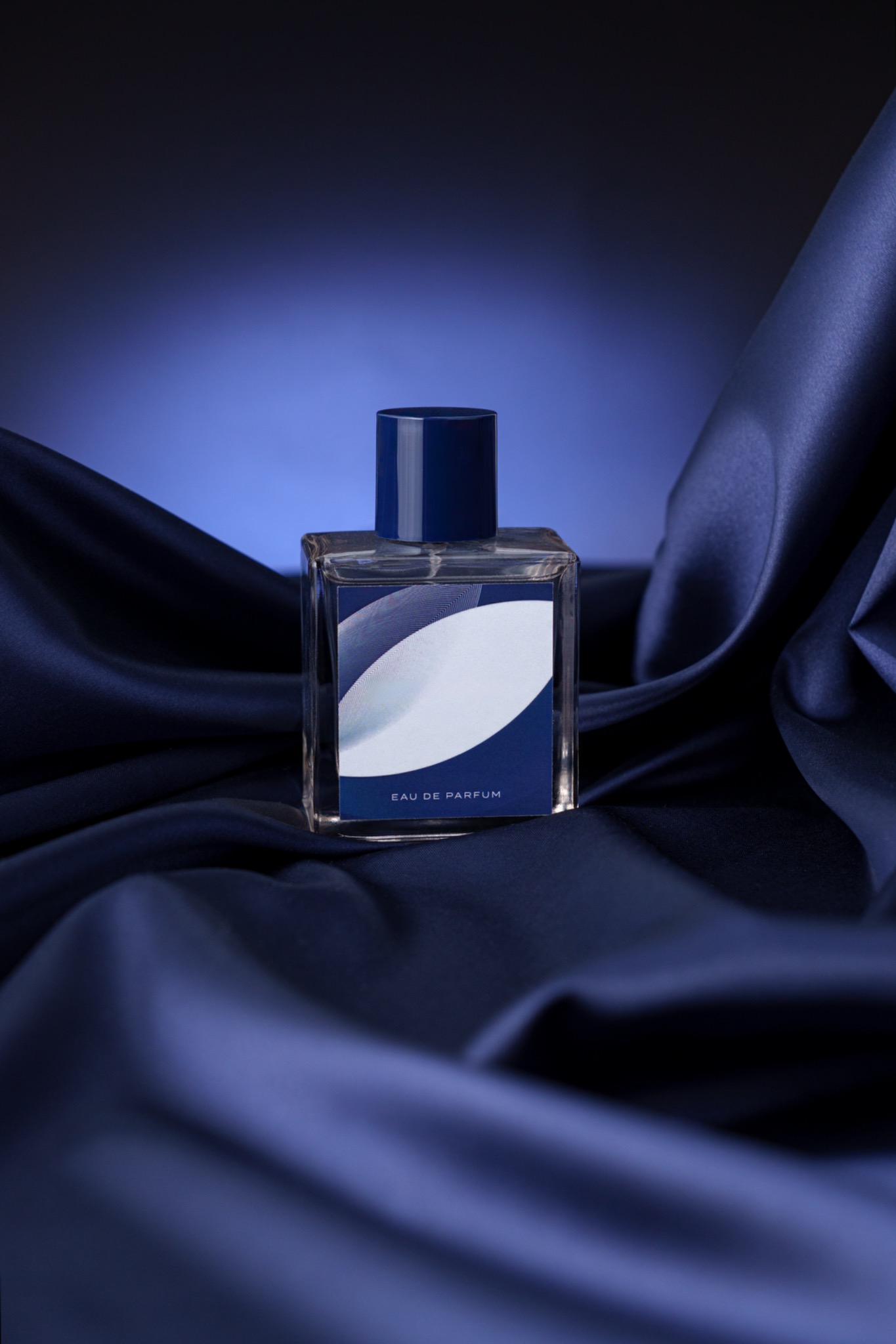 Kwadratowy flakon perfum Eau de Parfum z niebieską etykietą i zatyczką, ustawiony na miękkiej, ciemnoniebieskiej satynowej tkaninie z subtelnym oświetleniem.