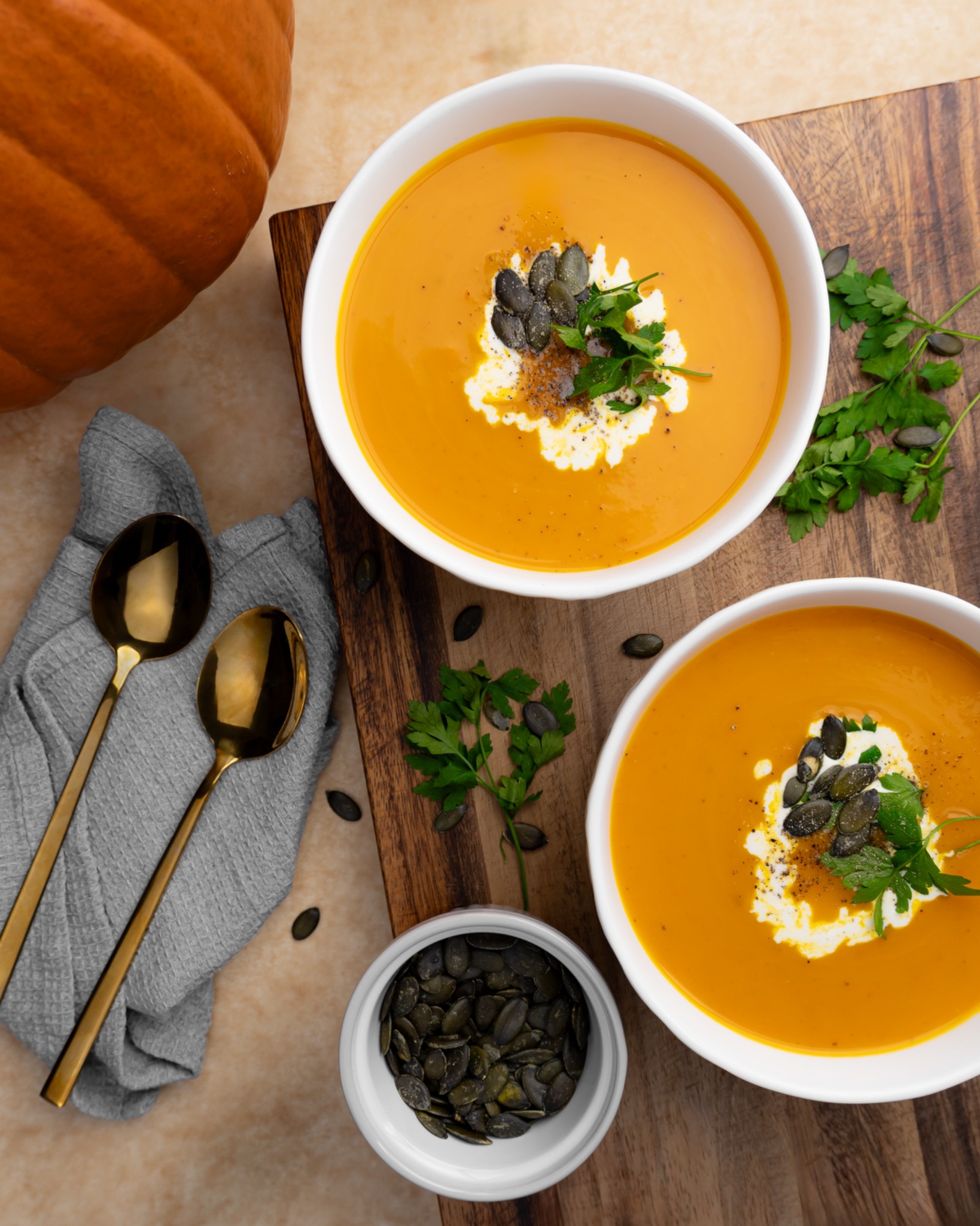 Dwa talerze kremowej zupy dyniowej, udekorowane kleksem śmietany, pestkami dyni i świeżą natką pietruszki, ułożone na drewnianej desce obok dyni, szarych ściereczek i złotych łyżek.