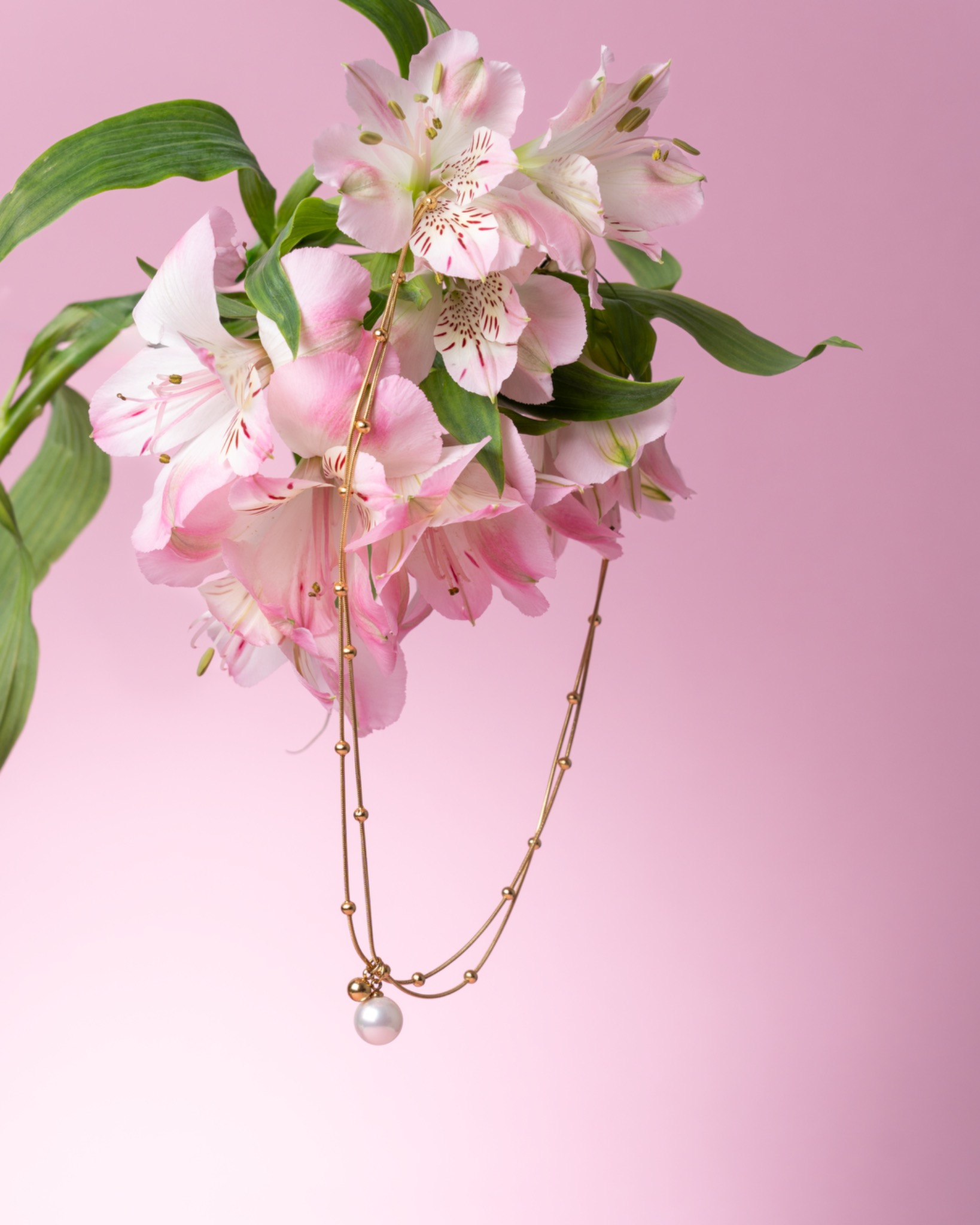 Delikatny złoty naszyjnik z perłą zwisa z bukietu różowych alstremerii na jednolitym różowym tle, prezentowany przez fotografa produktowego z Warszawy.
