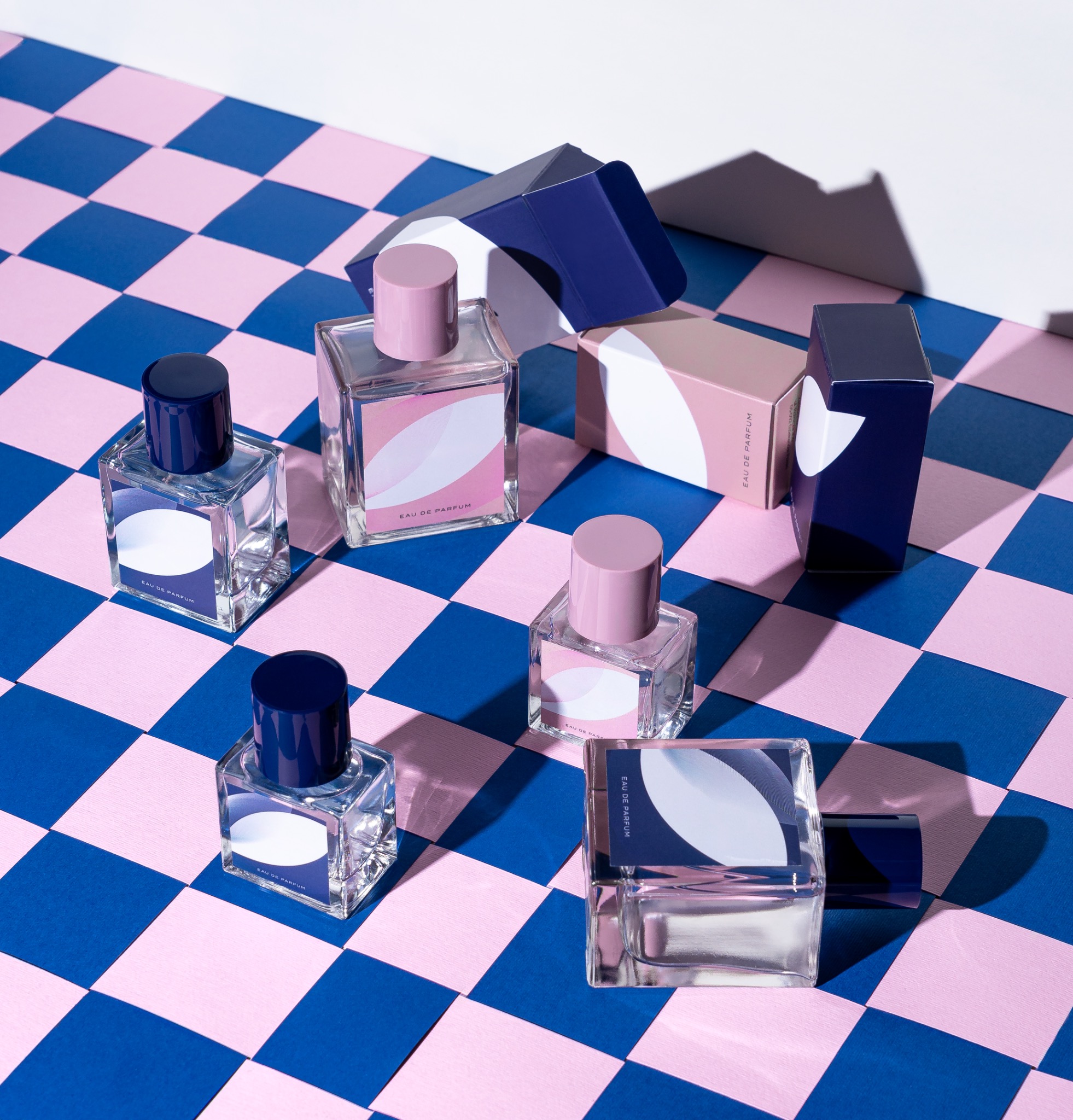 Kompozycja kilku flakonów perfum z minimalistycznymi etykietami na tle szachownicy w odcieniach różu i granatu, pudełka w podobnej kolorystyce w tle.