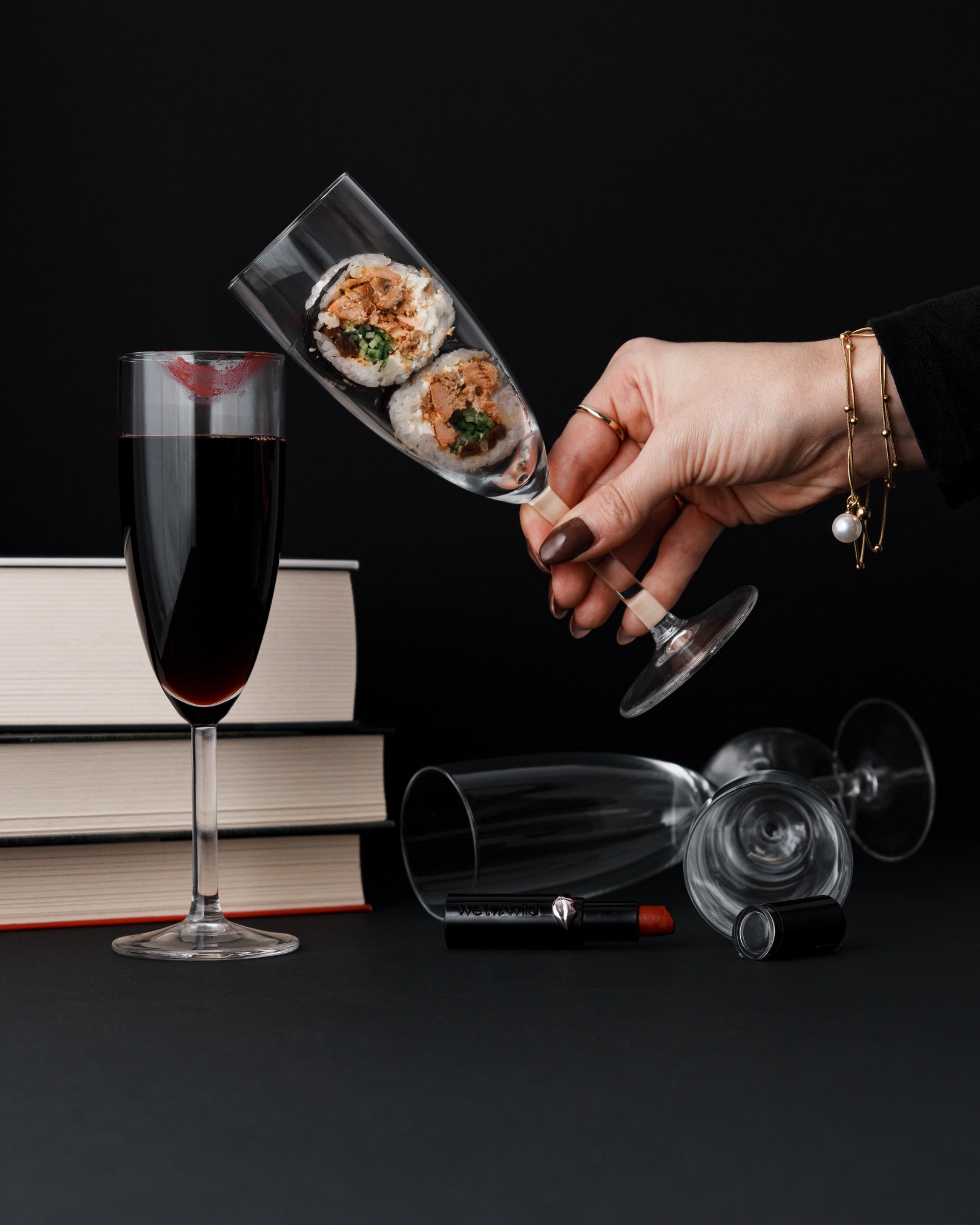 Dwa kawałki sushi w kieliszku do szampana trzymanym przez kobiecą dłoń z brązowymi paznokciami i złotą bransoletką, obok kieliszka z czerwonym winem i odciskiem szminki, leżącego obok stosu książek...