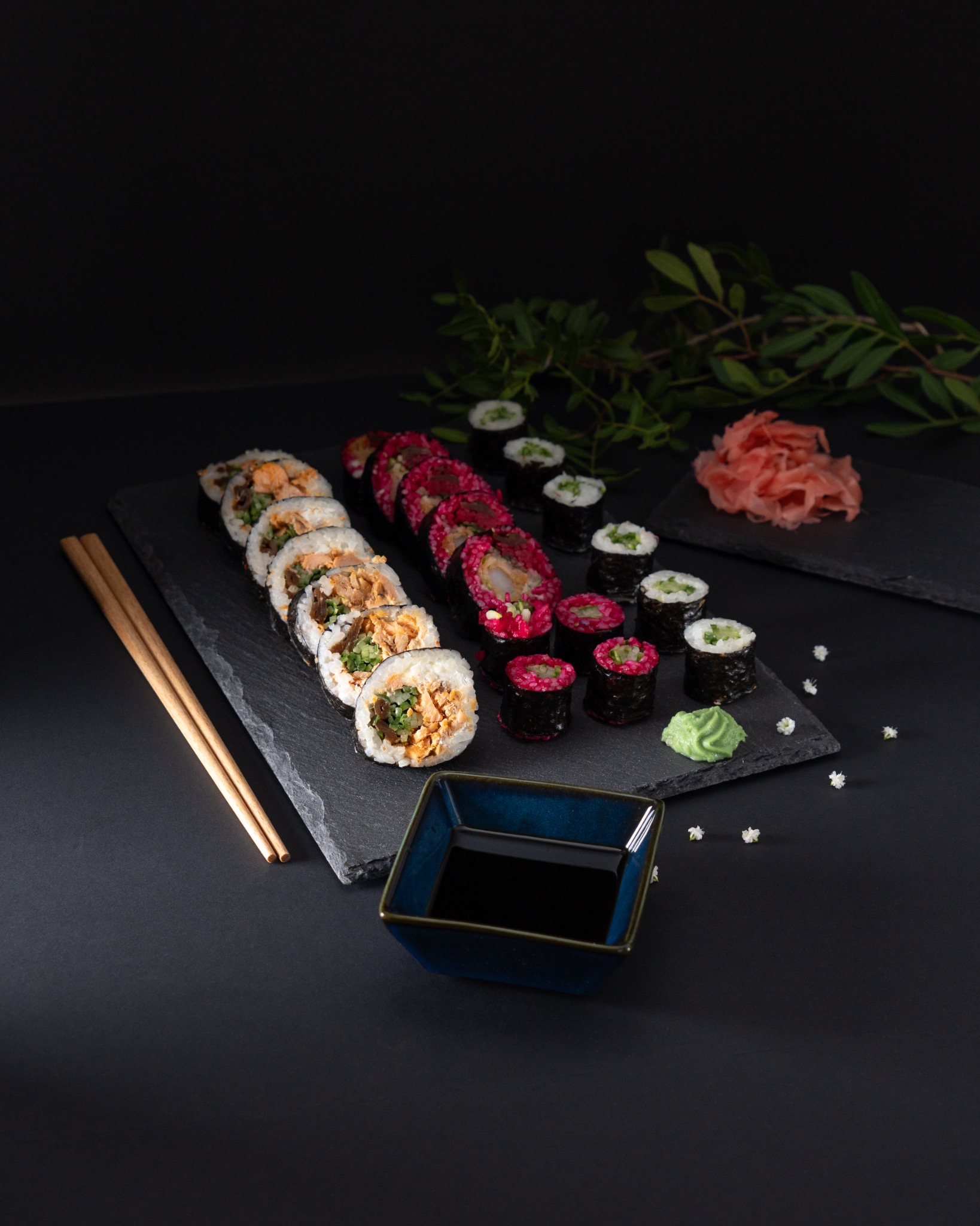 Profesjonalna fotografia produktowa: Zestaw sushi z różnymi rodzajami rolek, podany na czarnym łupku z sosem sojowym, wasabi i imbirem, w otoczeniu zielonych gałązek i drobnych białych kwiatów,...