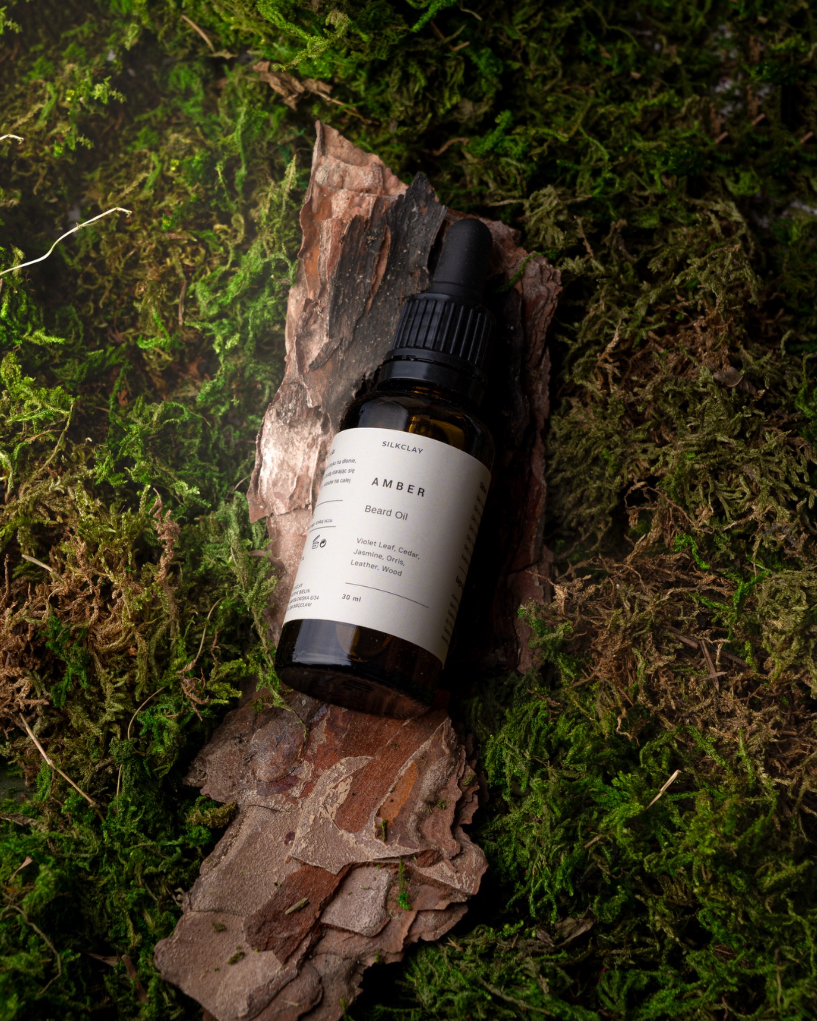 Butelka olejku do brody Amber marki Silkclay, umieszczona na korze drzewa, otoczona zielonym mchem.