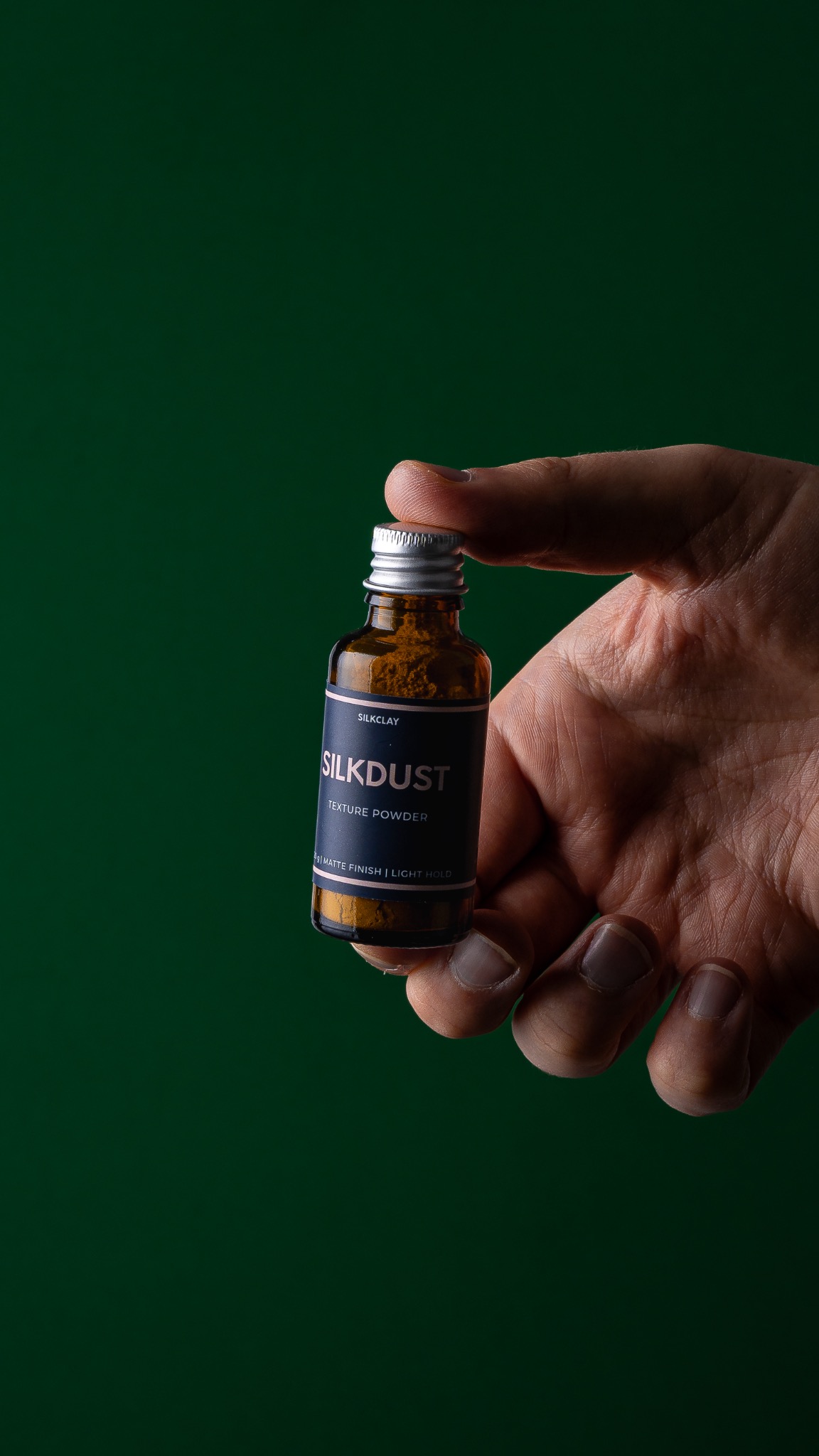 Dłoń trzymająca butelkę brązowego pudru tekstury Silkdust na ciemnozielonym tle.