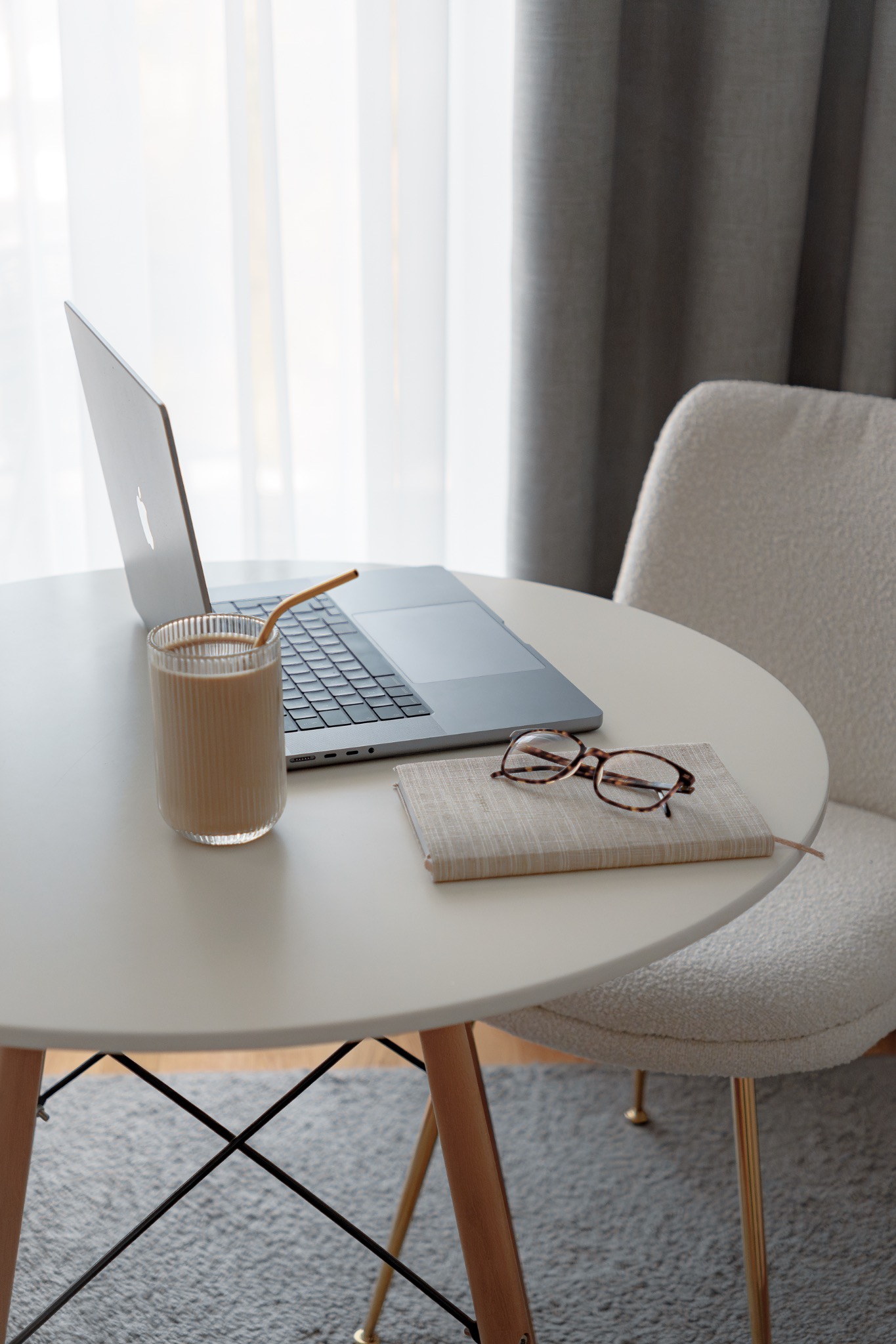 Minimalistyczne biurko z laptopem, szklanką kawy z brązową słomką, notatnikiem i okularami w brązowych oprawkach, na tle białej zasłony i szarej rolety, oświetlone naturalnym światłem.