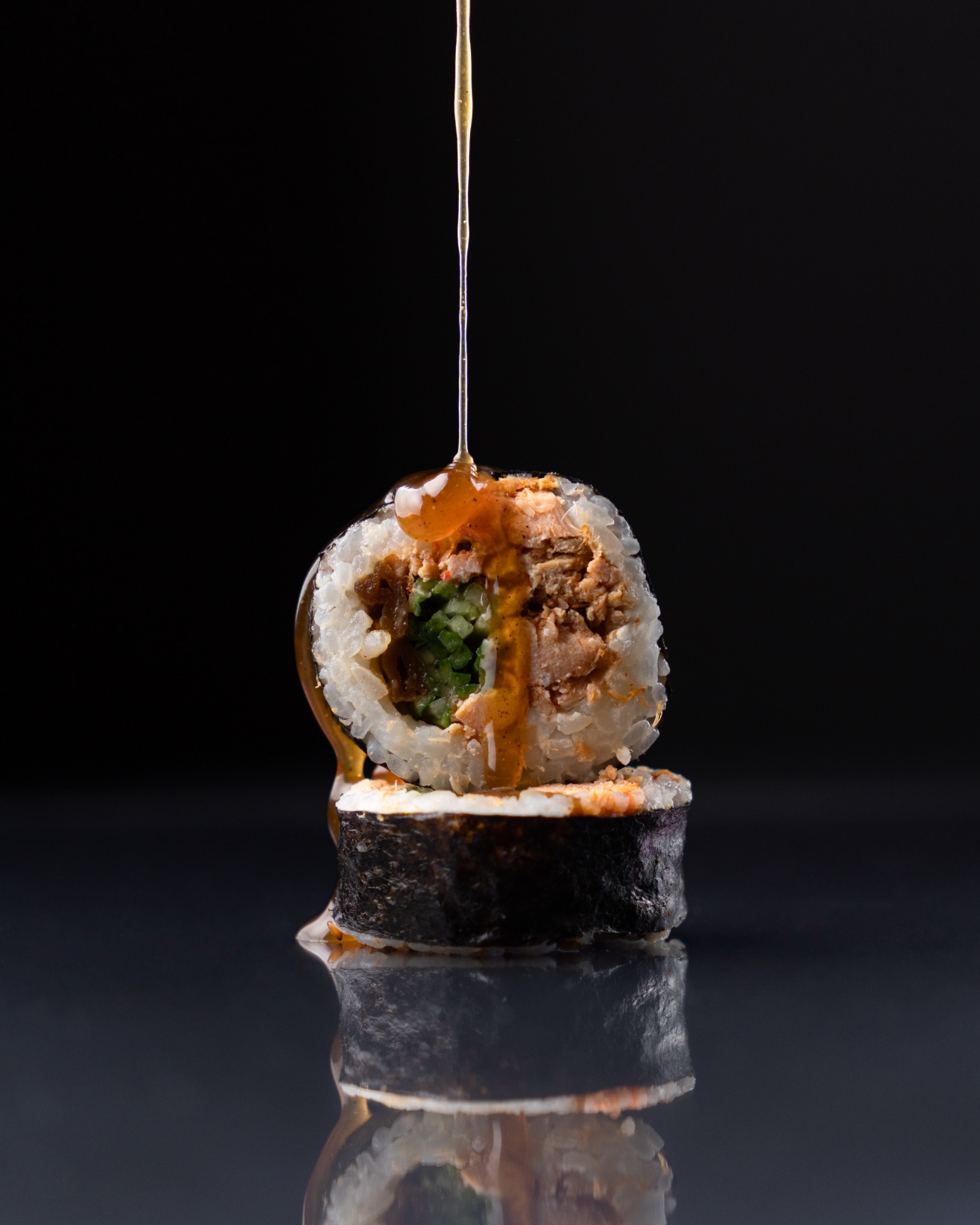 Dwa kawałki sushi ułożone jeden na drugim na błyszczącej powierzchni, polewane sosem z góry na czarnym tle.