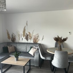 Grafic Wall - Jasne wnętrze salonu z szarym narożnikiem, okrągłym stołem z bukietem suszonych traw i naklejkami na ścianie imitującymi trawy pampasowe.