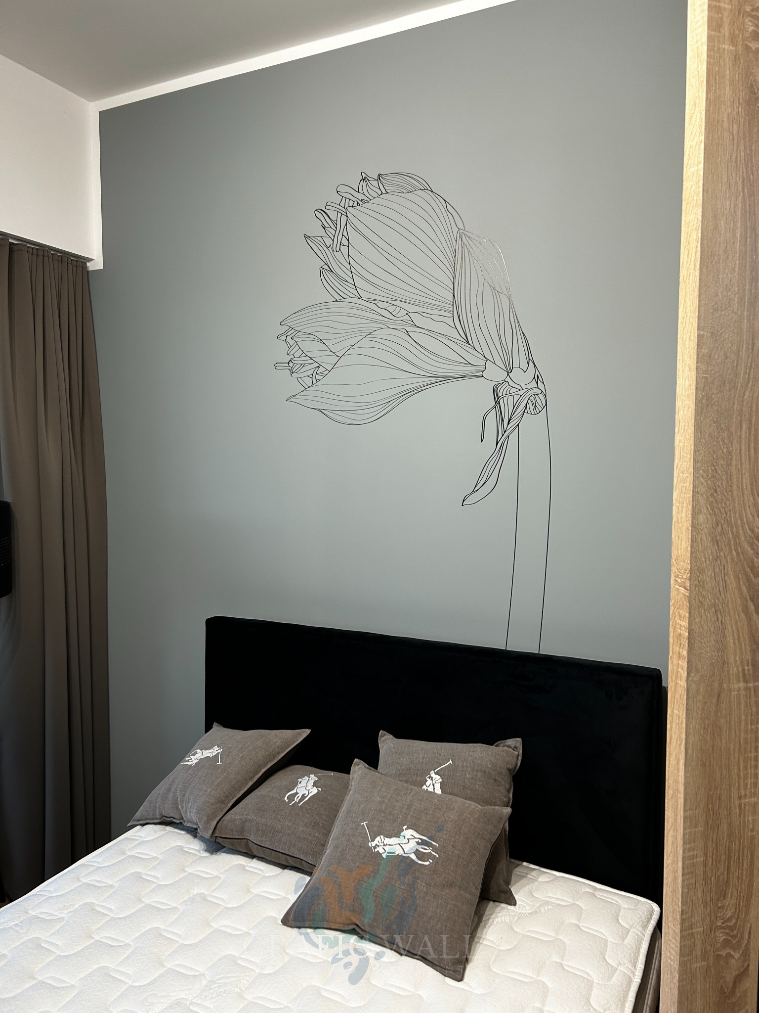 Sypialnia z szarą ścianą ozdobioną czarnym rysunkiem kwiatu, czarnym zagłówkiem łóżka i poduszkami z motywem jeźdźca polo.