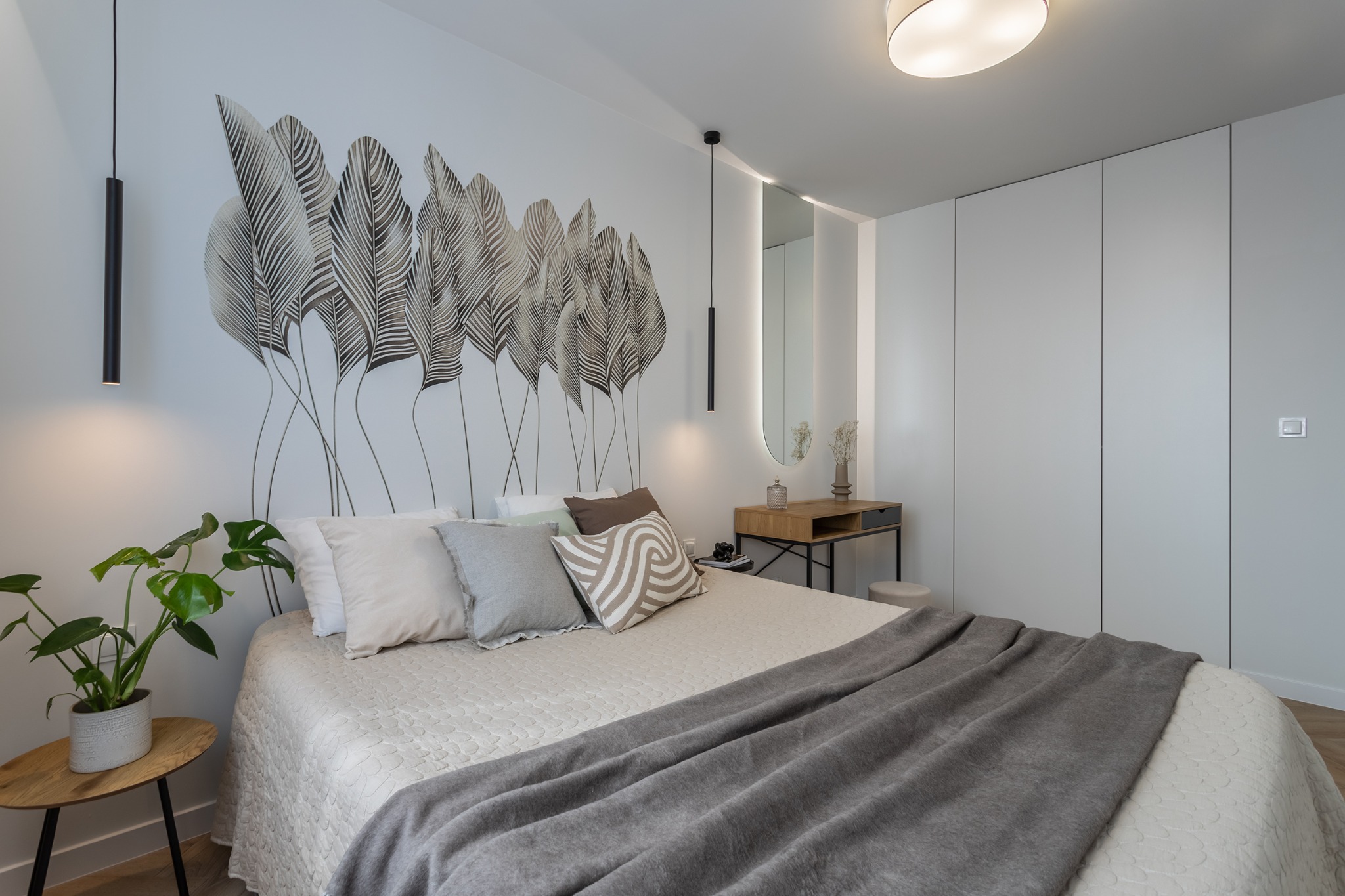 Jasna sypialnia z łóżkiem, stolikiem nocnym z rośliną Monstera, toaletką z owalnym lustrem podświetlonym LED i wzorem liści na ścianie za wezgłowiem.