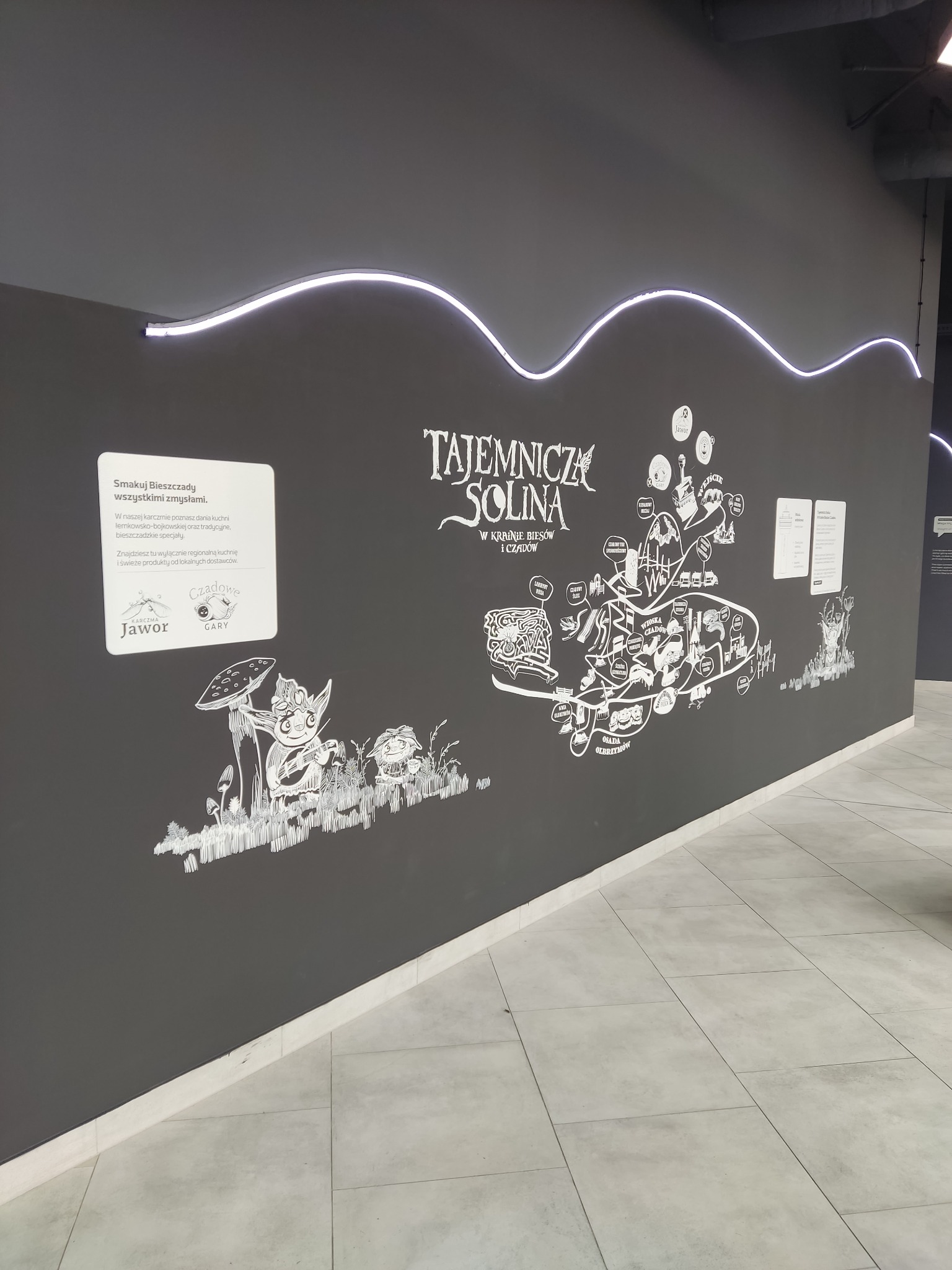 Czarna ściana z białymi rysunkami i napisami informacyjnymi w lokalu gastronomicznym, oświetlona ledowym paskiem, prezentująca mapę atrakcji regionu Solina.