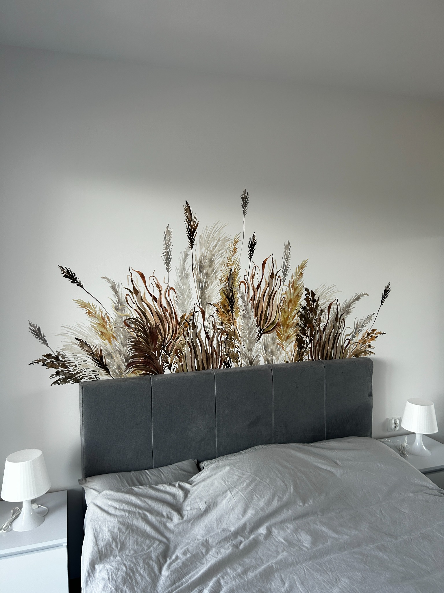 Sypialnia z szarym łóżkiem i naklejką ścienną przedstawiającą trawy pampasowe nad wezgłowiem, białe szafki nocne z lampkami po bokach.