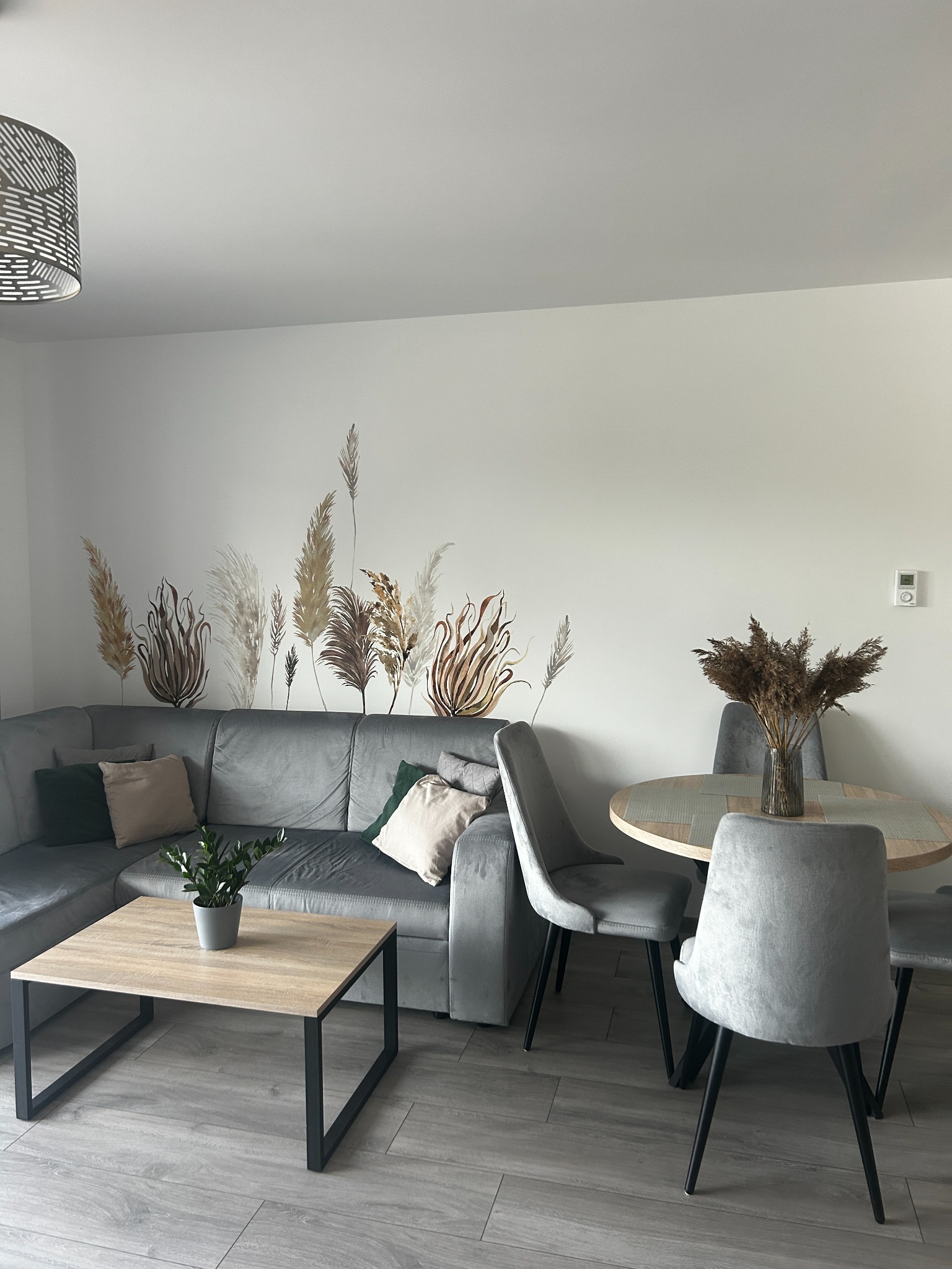 Jasne wnętrze salonu z szarym narożnikiem, okrągłym stołem z bukietem suszonych traw i naklejkami na ścianie imitującymi trawy pampasowe.