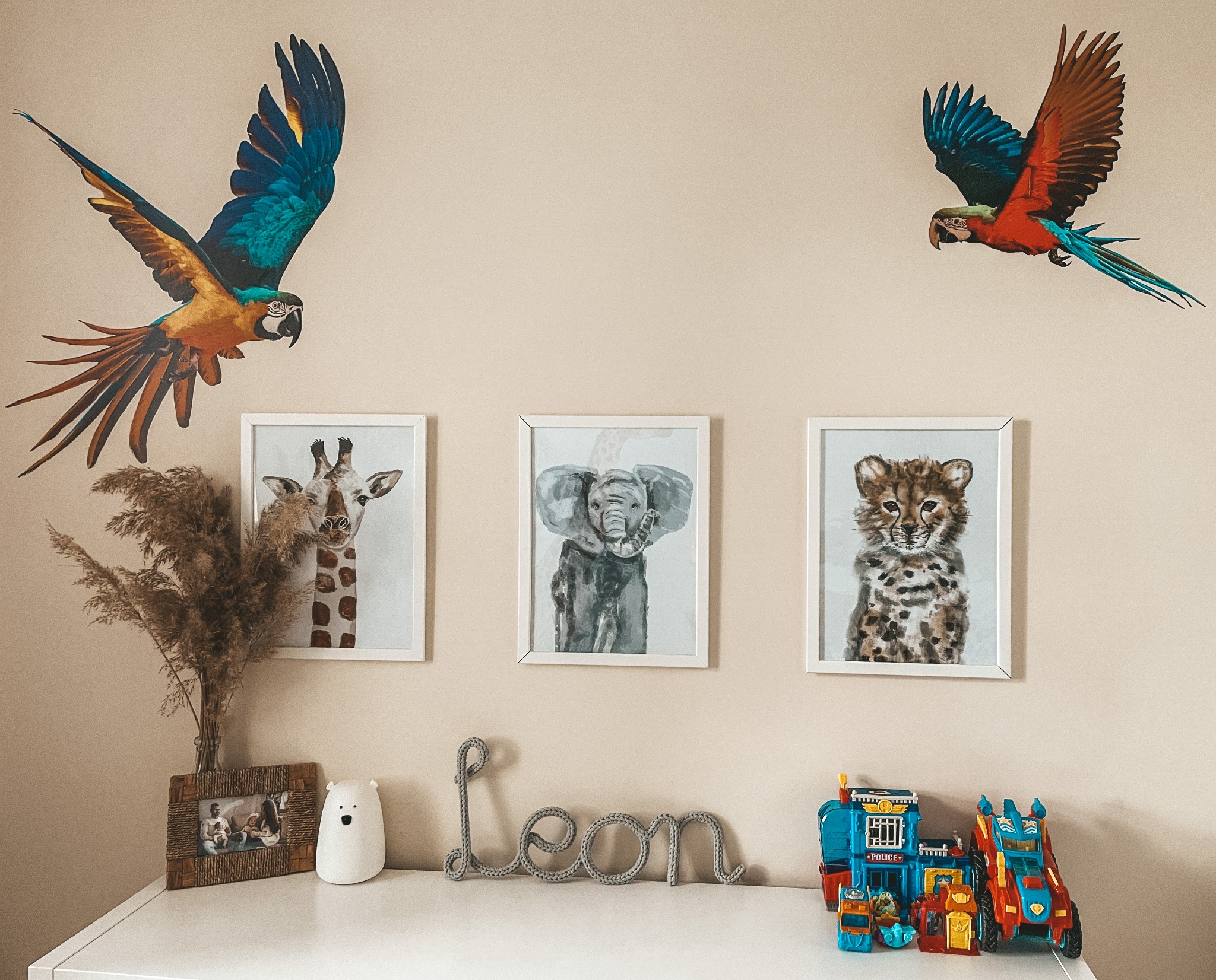 Pokój dziecięcy z dekoracjami ściennymi: naklejki z papugami oraz oprawione grafiki ze zwierzętami (żyrafa, słoń, gepard), dekoracyjny napis 'Leon' i zabawki.