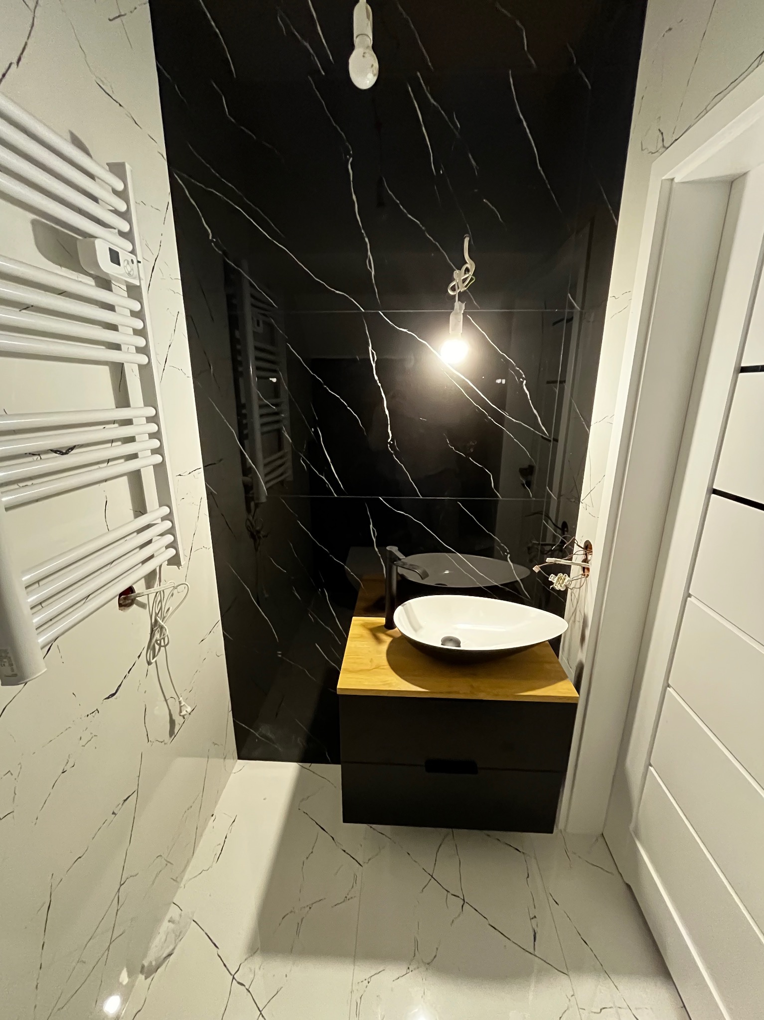 Nowoczesna łazienka z czarno-białymi marmurkowymi płytkami, wiszącą białą umywalką na drewnianym blacie i czarną szafką pod umywalką, oraz grzejnik drabinkowy.