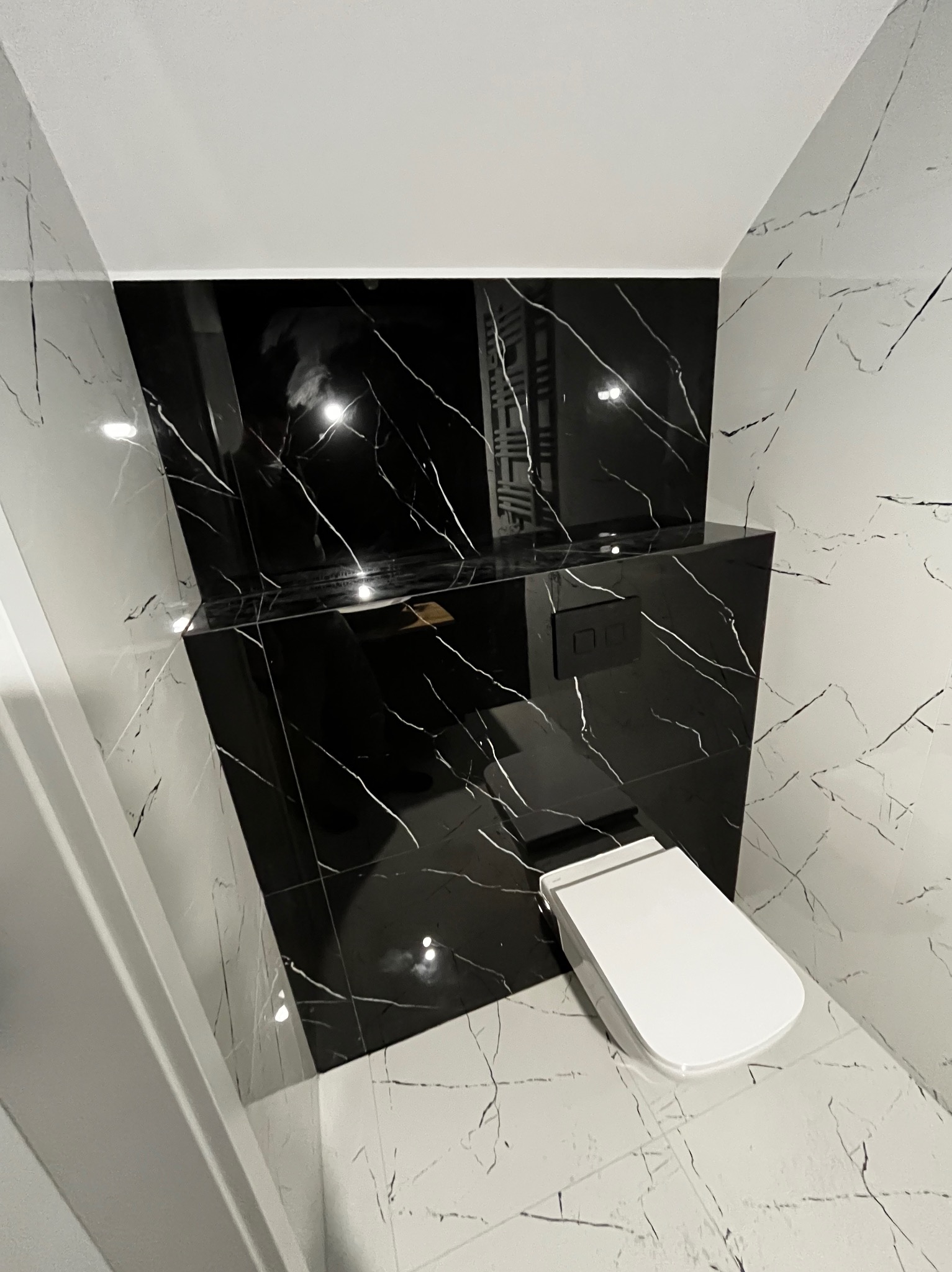 Nowoczesna łazienka z czarno-białymi płytkami imitującymi marmur, wisząca toaleta i zabudowa z czarnych płytek.
