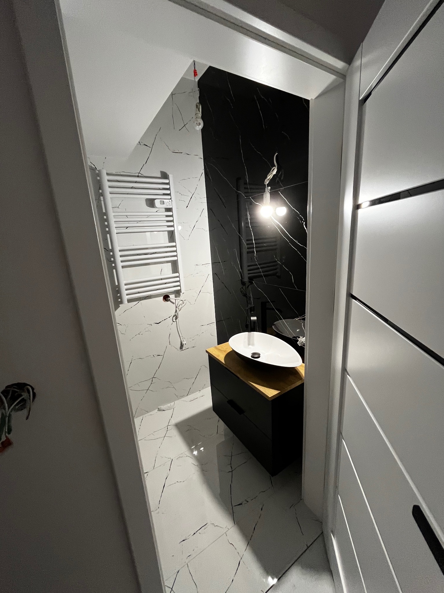 Nowoczesna łazienka z czarno-białymi marmurkowymi płytkami, wiszącą szafką z drewnianym blatem i owalną umywalką, widziana zza częściowo otwartych drzwi.