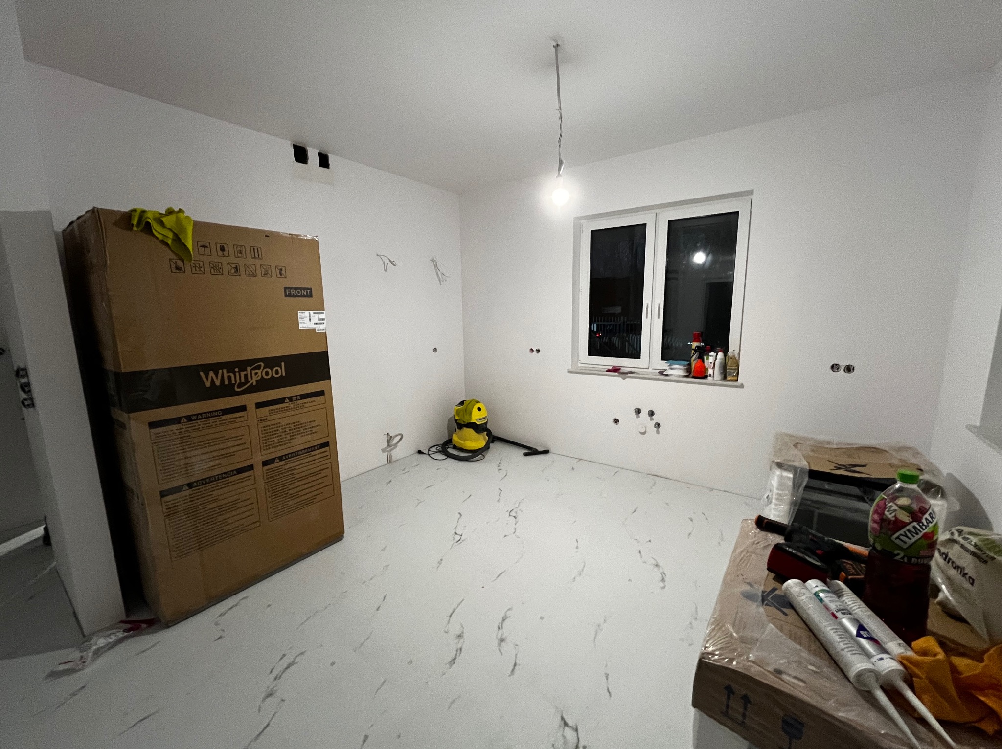 Wnętrze w trakcie przygotowań do prac wykończeniowych: karton z nowym sprzętem Whirlpool, odkurzacz, materiały budowlane i narzędzia na podłodze z białego marmuru, surowe ściany z punktami...