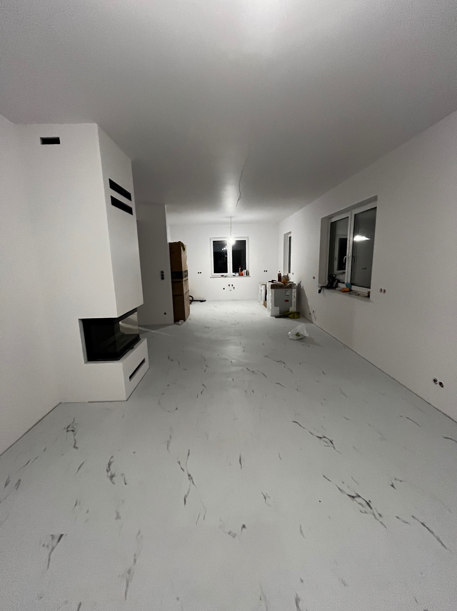 Wnętrze pokoju w trakcie prac wykończeniowych, z białymi ścianami, podłogą imitującą marmur, nowoczesnym kominkiem, oknami i widocznymi śladami po gipsowaniu.